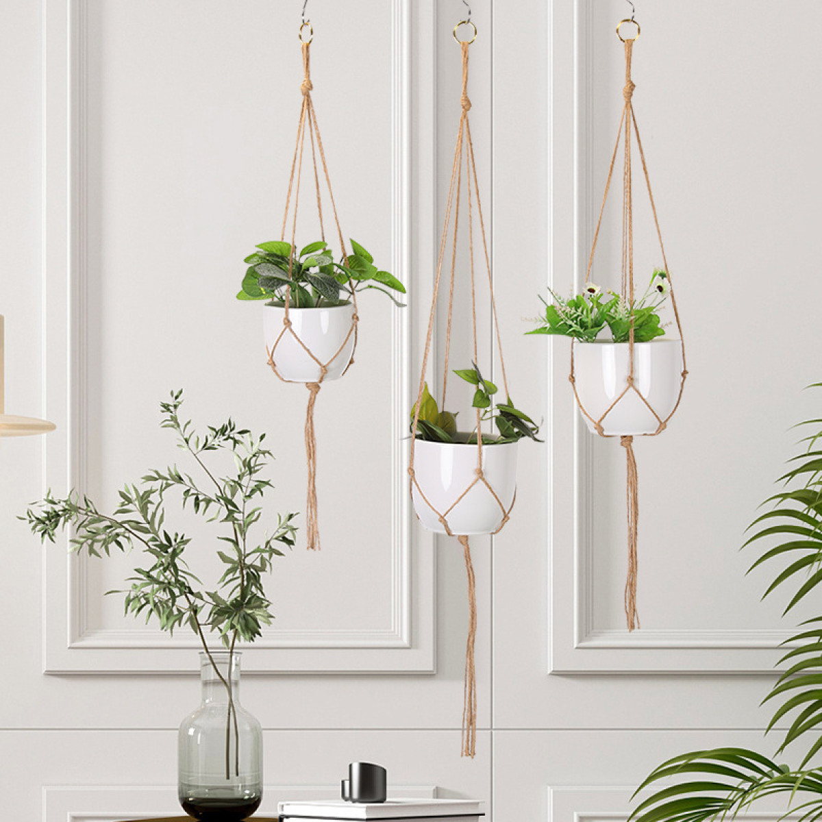 Drei hängende weiße Blumentöpfe mit grünen Pflanzen. Eine Vase mit grünen Blättern steht im Vordergrund. Der Hintergrund ist eine weiße Wand.