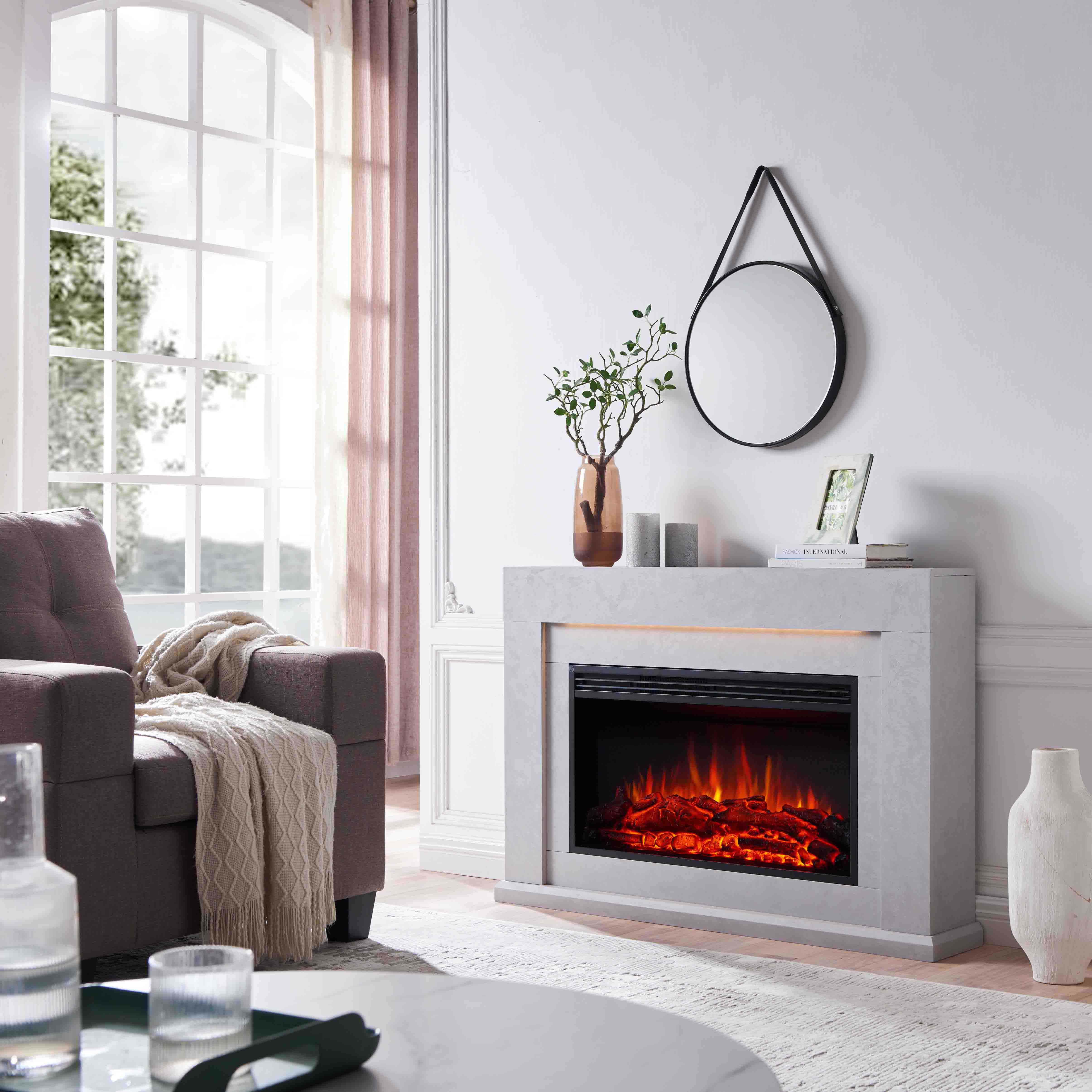 Ein Wohnzimmer mit Kamin, Sofa und großem Fenster. Über dem Kamin hängt ein Spiegel, daneben steht eine Vase.