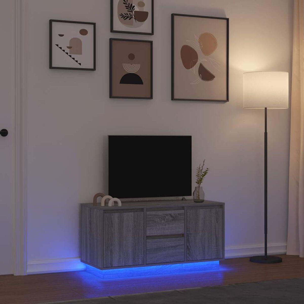 Ein Fernseher auf einem grauen Schrank mit blauem Unterleuchten, gerahmte Kunst an der Wand und eine Stehlampe. Eine Vase mit Pflanzen steht auf dem Schrank.