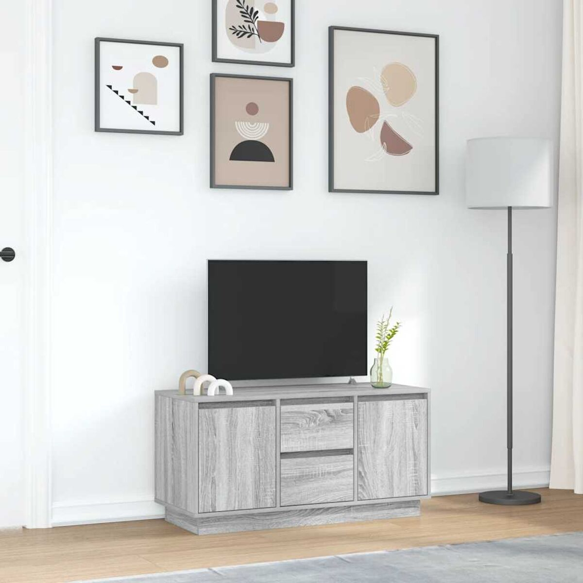 Ein Wohnzimmer mit einem Fernseher auf einem grauen Schrank. Oben sind gerahmte Bilder. Rechts steht eine Lampe.