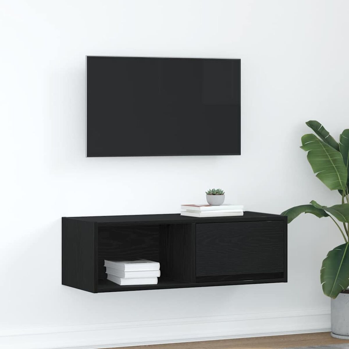 Ein schwarzer Fernseher und ein schwarzer Schrank sind an einer weißen Wand montiert. Bücher und eine kleine Pflanze stehen auf dem Schrank. Rechts steht eine Topfpflanze.