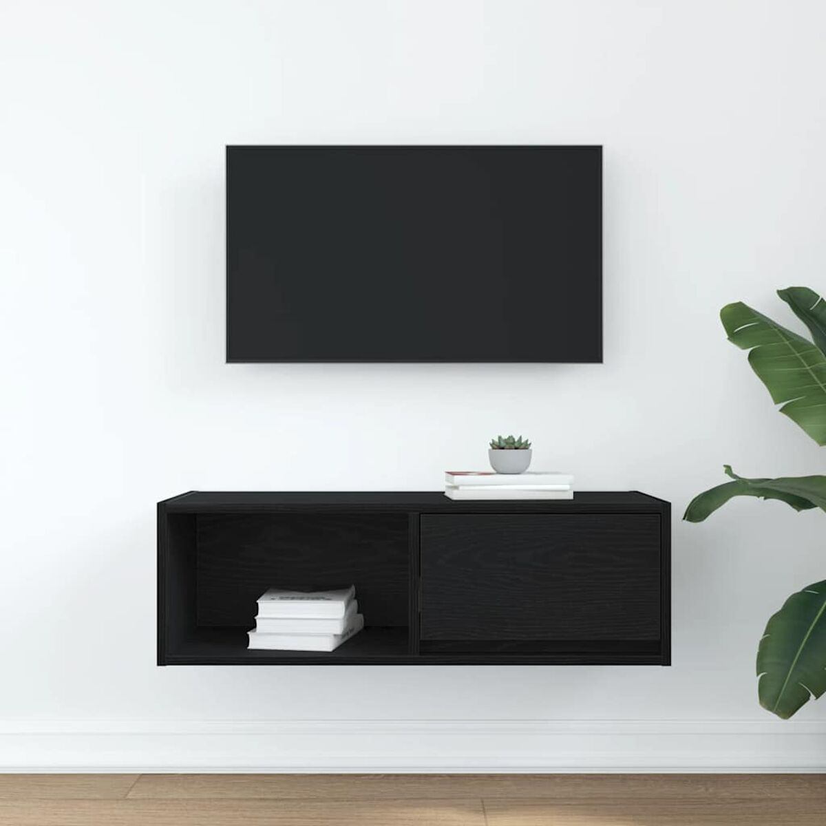 Ein schwarzer Fernseher über einem schwarzen Schrank mit Büchern und einer Pflanze. Eine große grüne Pflanze rechts. Die Wand ist weiß.