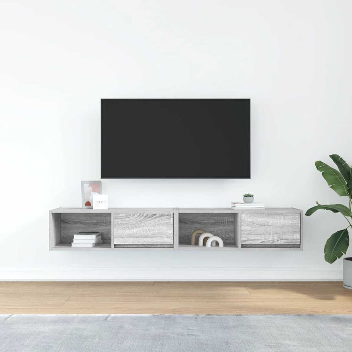 Ein Fernseher hängt über einem grauen, an der Wand befestigten Schrank mit Büchern, Deko und einer Pflanze. Ein Teppich und eine Topfpflanze sind ebenfalls vorhanden.