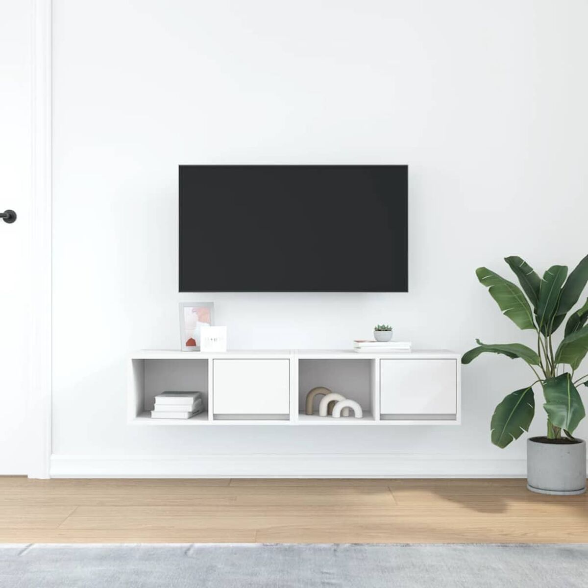 Ein Raum mit einem Fernseher an einer weißen Wand. Unter dem Fernseher befindet sich ein weißes Regal mit Dekoration. Rechts steht eine Topfpflanze.