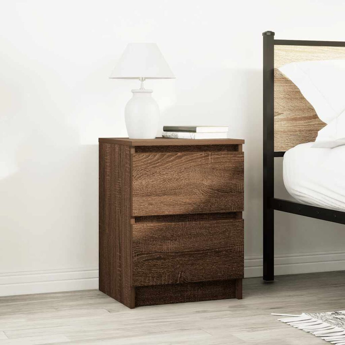 Ein Nachttisch aus Holz mit Lampe und Büchern neben einem Bett. Der Nachttisch hat zwei Schubladen. Dahinter befindet sich eine weiße Wand.