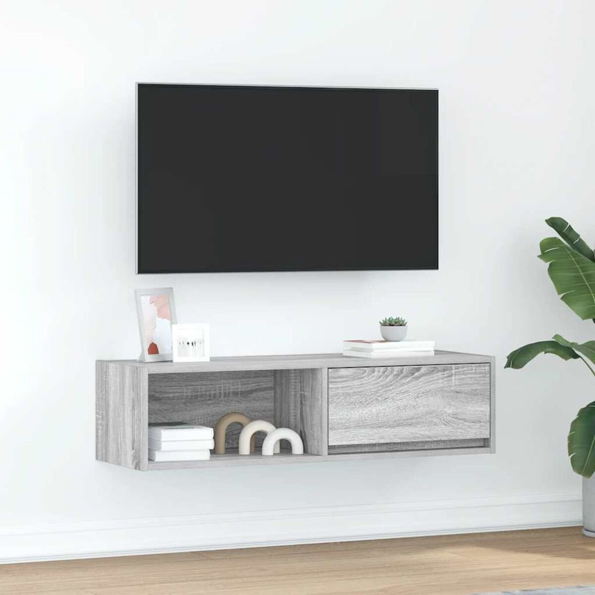 Ein Fernseher über einem grauen Schrank mit Dekoration. Rechts ist eine Pflanze. Die Wand ist weiß.