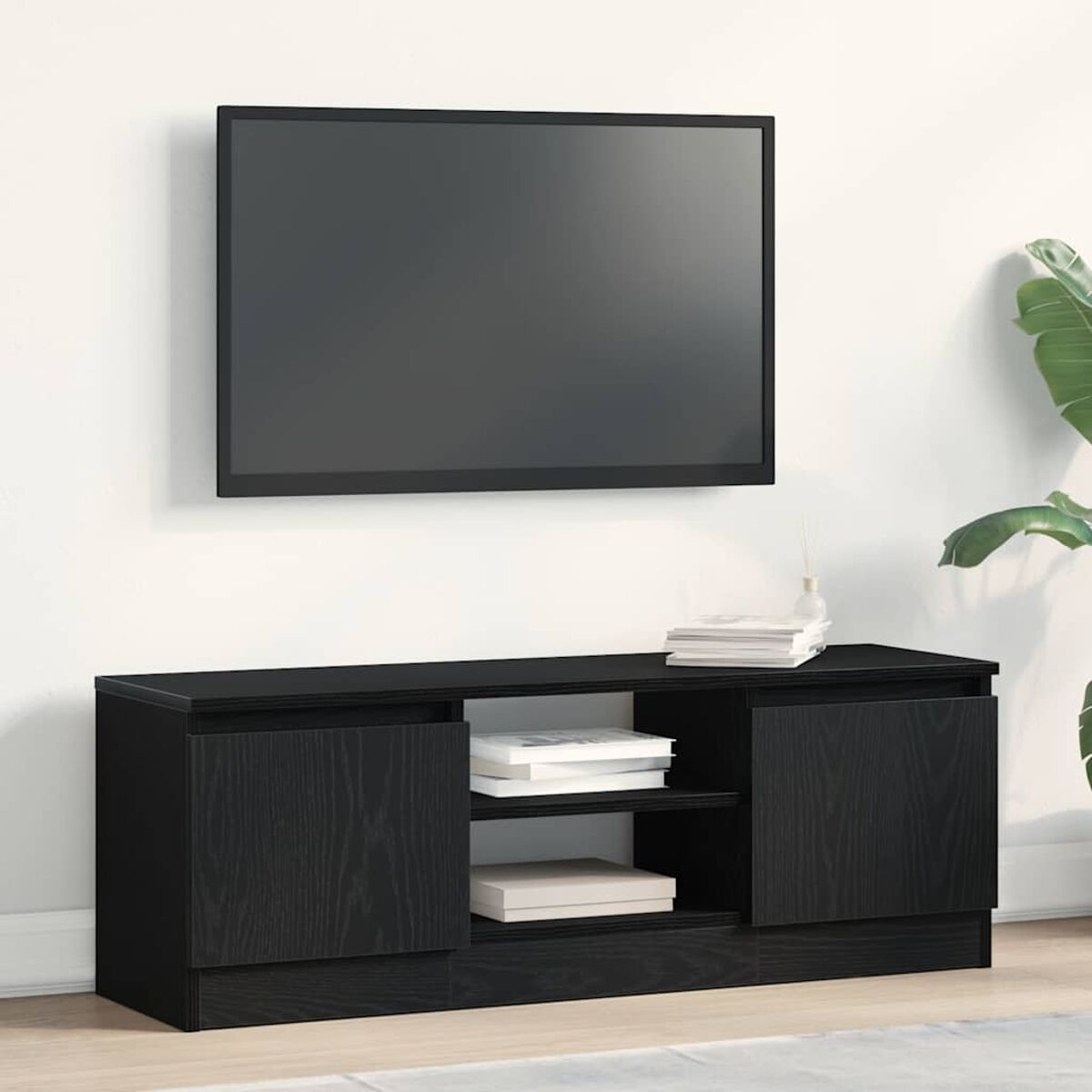 Ein schwarzer Fernseher ist über einer schwarzen Medienkonsole montiert. Bücher und eine Vase stehen auf der Konsole. Rechts ist eine Pflanze.