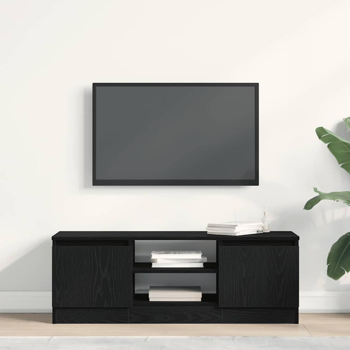 Unter einem schwarzen Fernseher befindet sich ein schwarzer TV-Ständer. Bücher und eine Pflanze stehen daneben an einer weißen Wand.