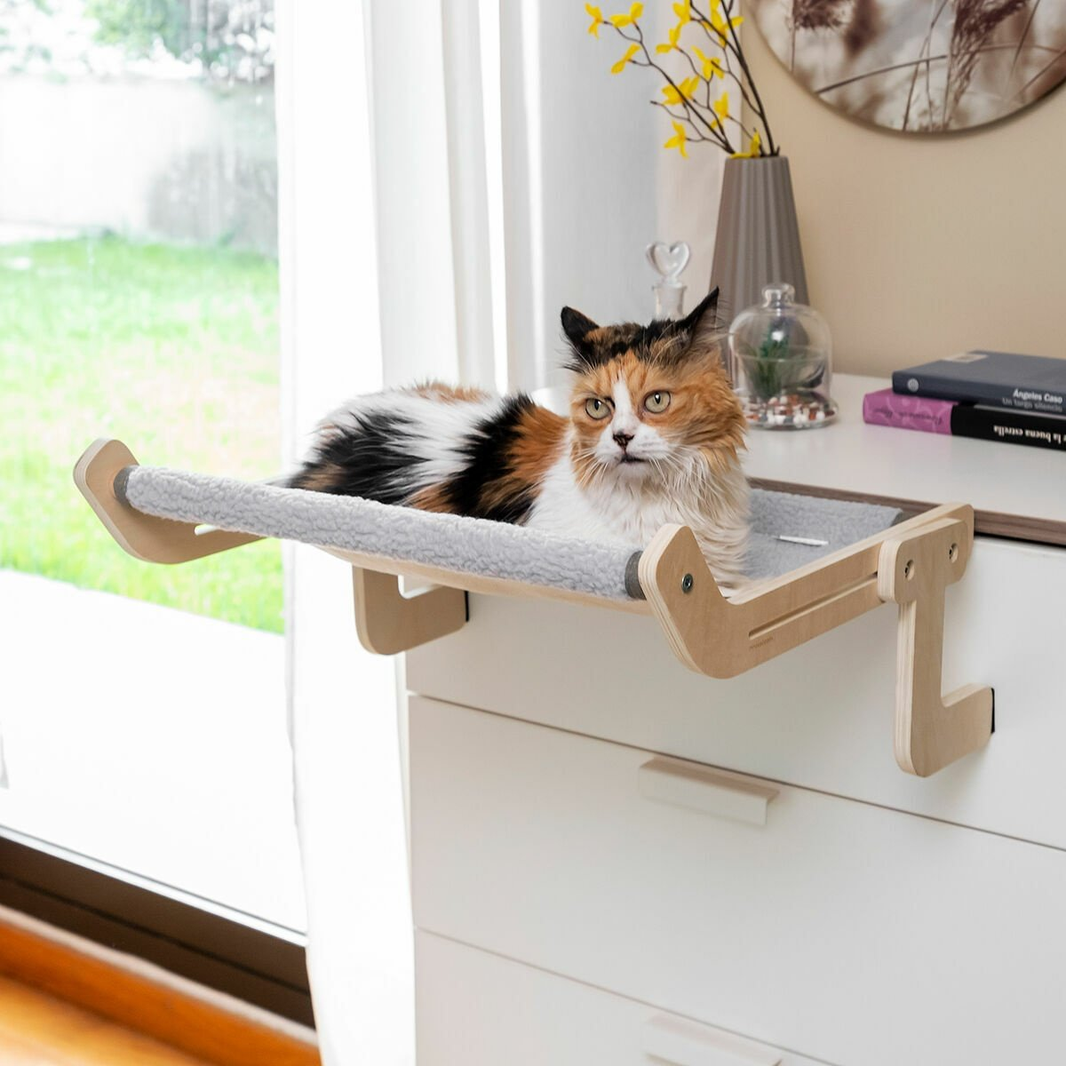 Un gatto calico riposa su un lettino di legno per gatti attaccato a un comò bianco. Sullo sfondo sono visibili una finestra e delle decorazioni.