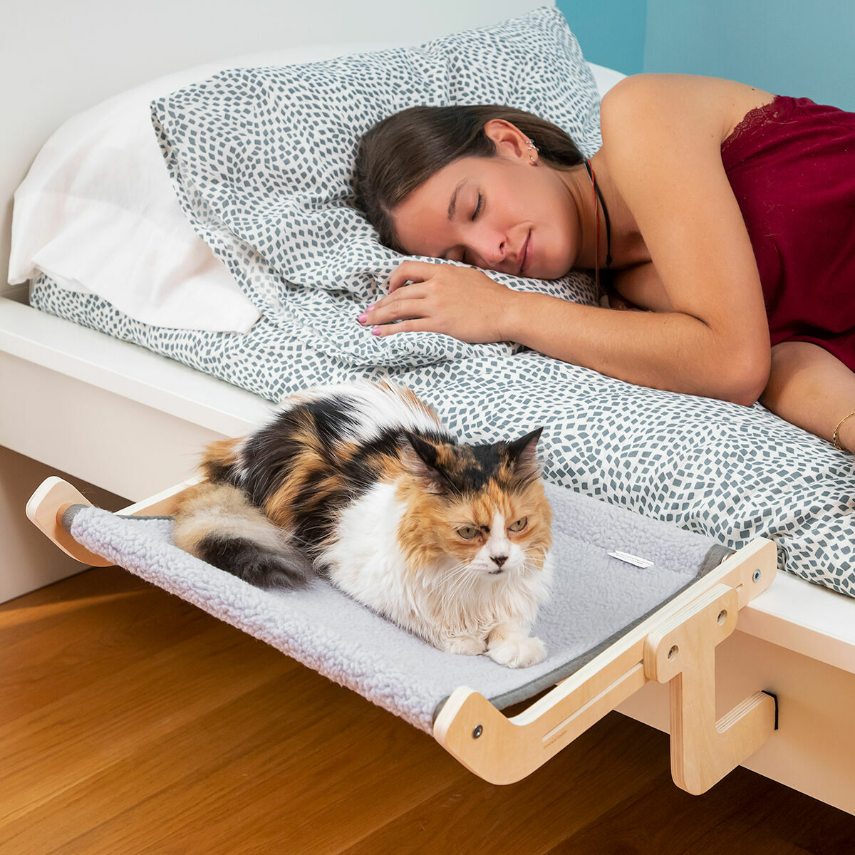 Una donna dorme a letto. Un gatto riposa su un'amaca per gatti montata sul letto. Il gatto è calico, il letto ha biancheria da letto fantasia e struttura in legno.