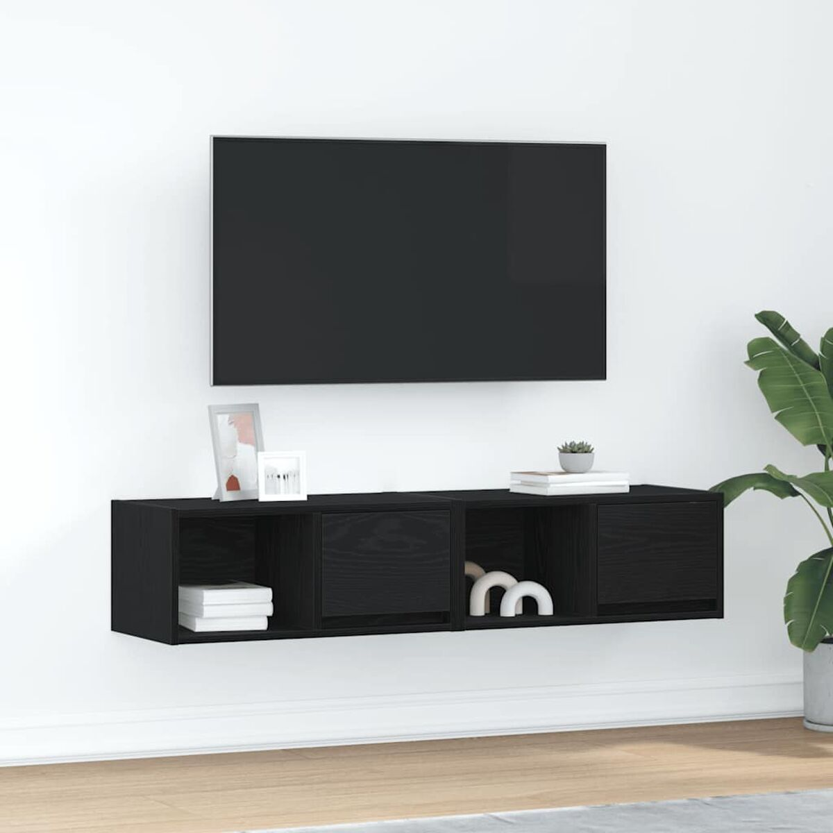 Schwarzes, an der Wand befestigtes TV-Möbel mit einem Fernseher darüber. Es hat Stauraum und Dekorationsartikel. Rechts steht eine Pflanze.