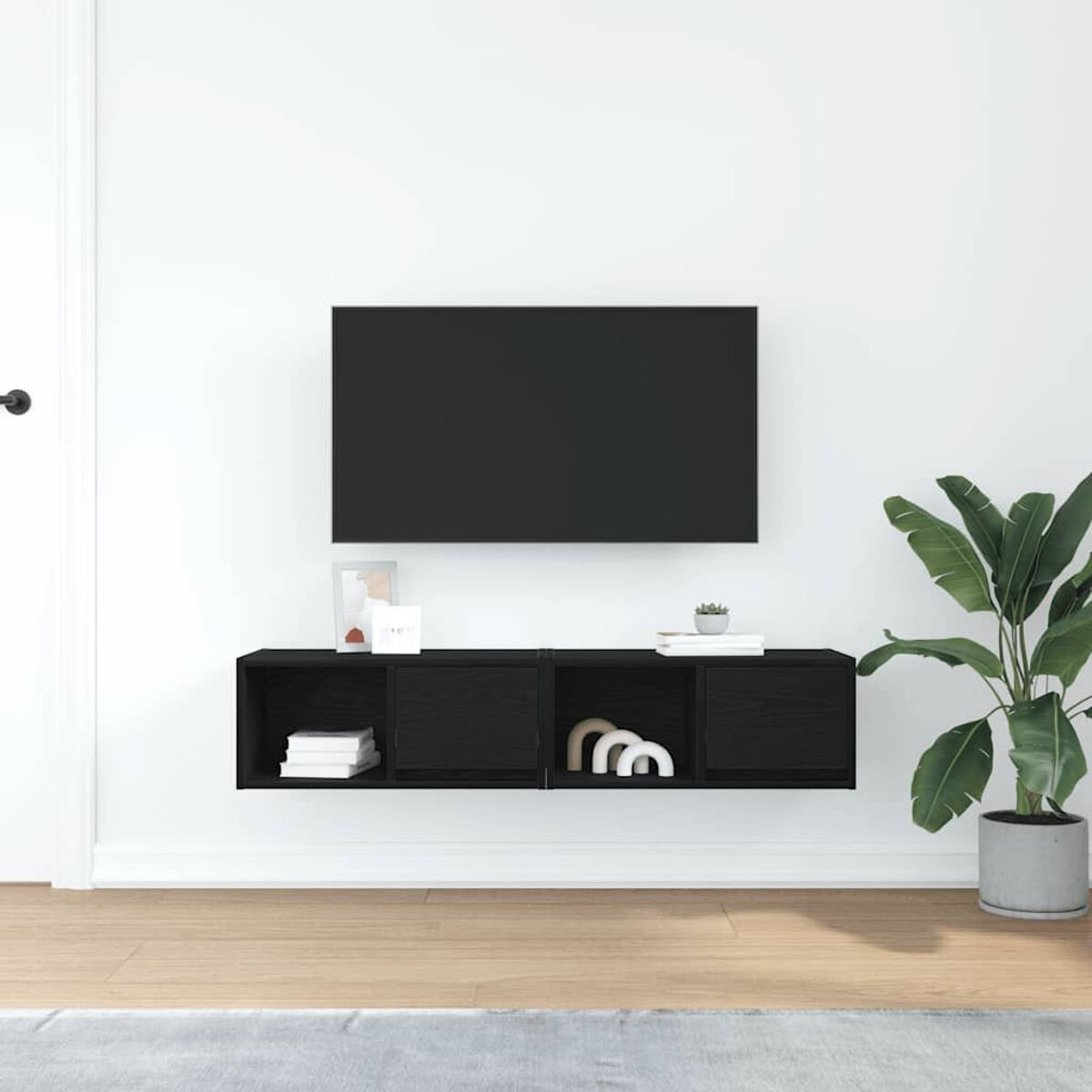 Ein Wohnzimmer mit Wand-TV, schwarzem Stauraum, einer Pflanze und weißer Wand. Der Boden ist aus Holz.