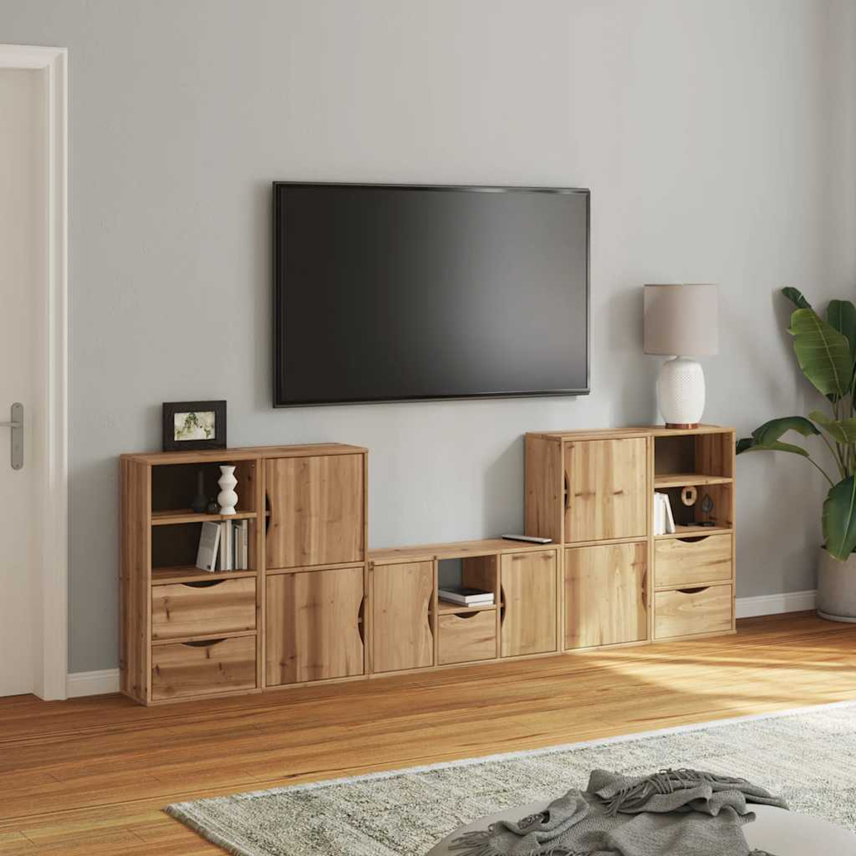 Ein Wohnzimmer mit Holzmöbeln, einem Fernseher, einer Lampe und einer Pflanze. Links ist eine Tür. Ein Teppich und Holzboden sind sichtbar.