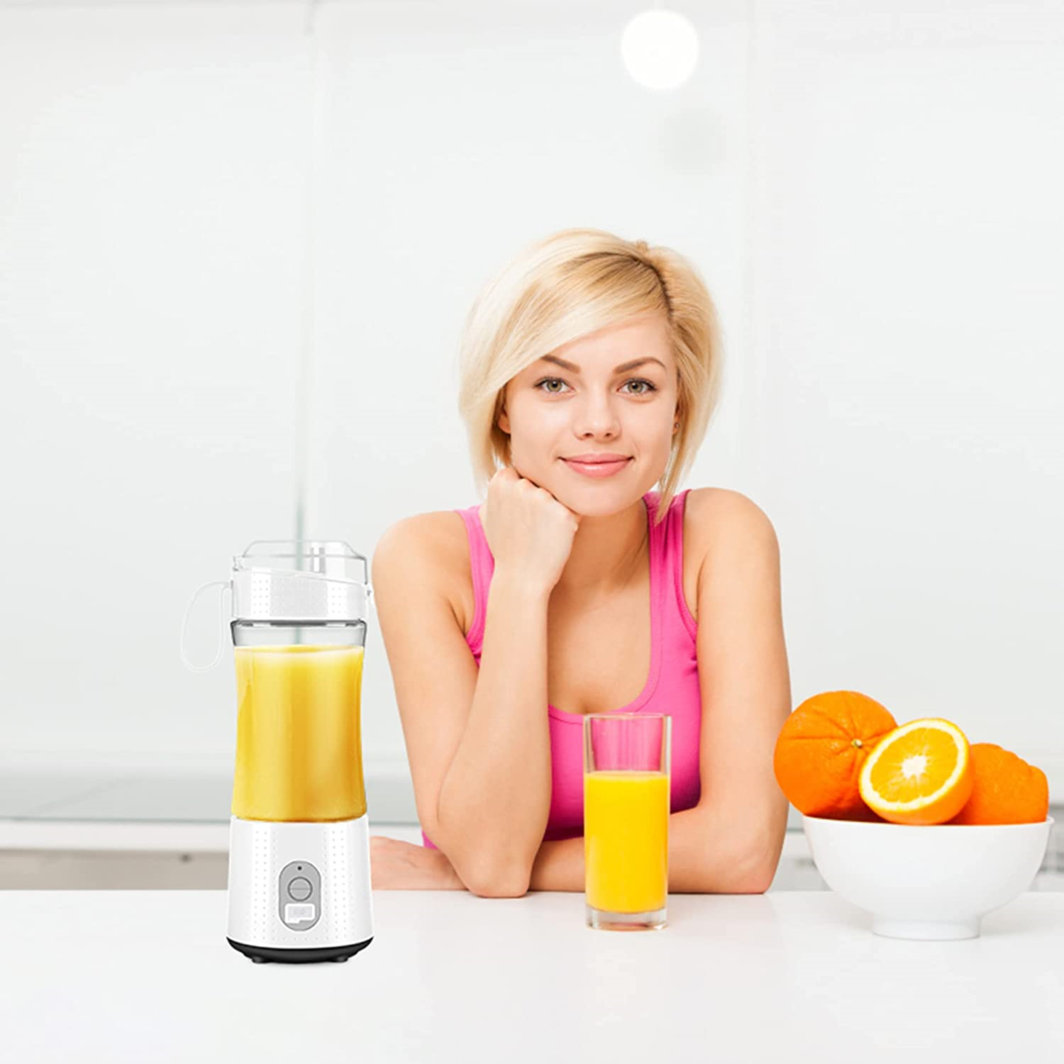 Eine Frau mit blonden Haaren sitzt an einem Tisch. Ein Mixer und Saft stehen auf dem Tisch mit Orangen in einer Schüssel.