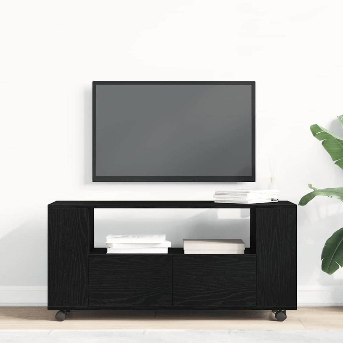Ein schwarzer TV-Ständer mit einem Fernseher darüber, weiße Wand, Bücher auf dem Ständer und eine Pflanze in der rechten Ecke.