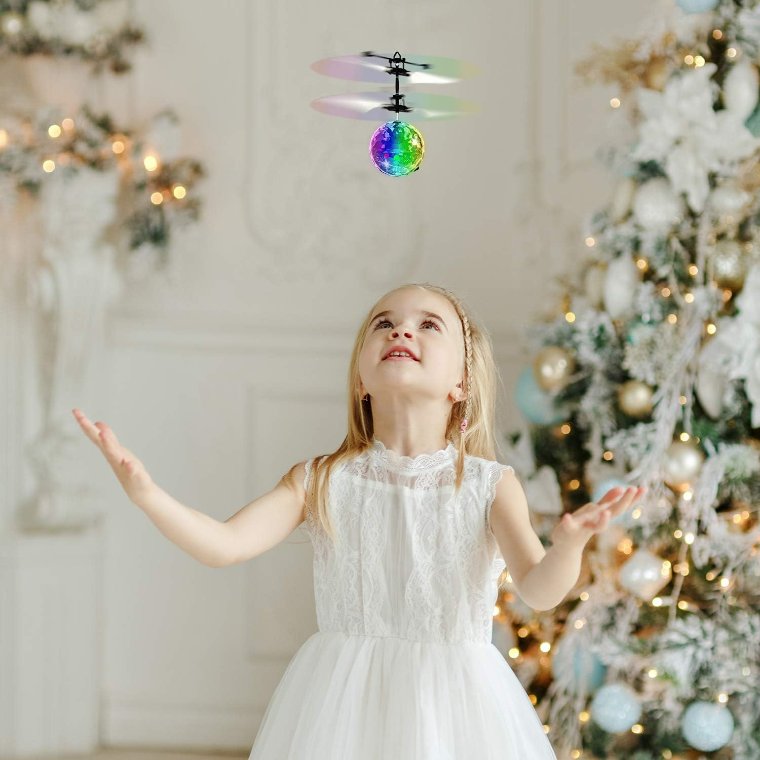 Ein junges Mädchen in einem weißen Kleid blickt auf ein fliegendes Spielzeug. Im Hintergrund ist ein Weihnachtsbaum.