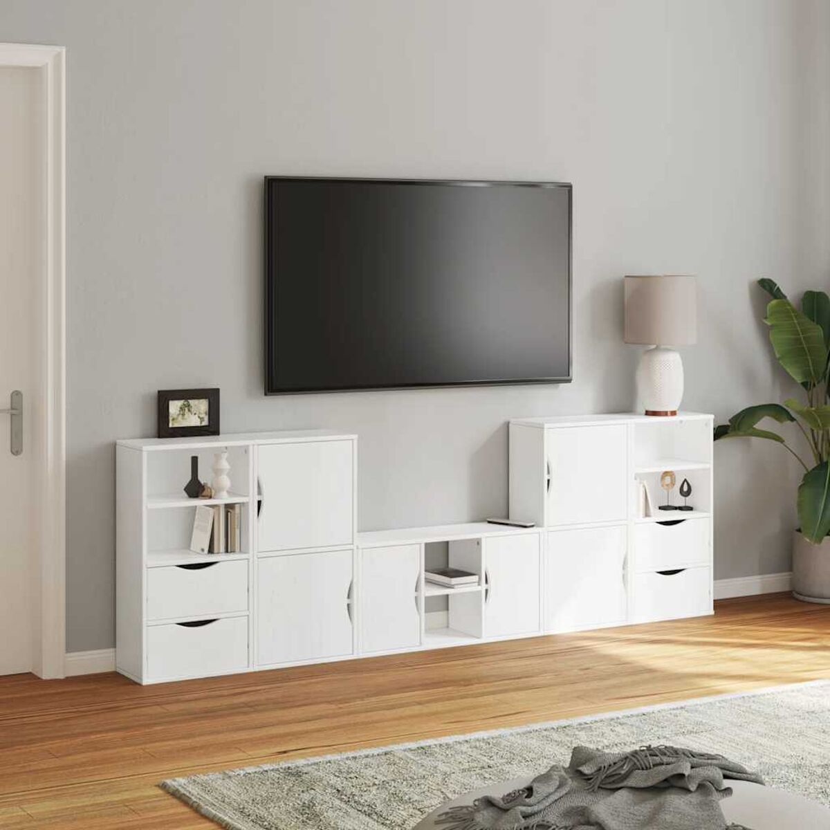 Wohnzimmer mit weißen Schränken, Fernseher, Lampe und Pflanze. Holzboden und ein Teppich im Vordergrund. Eine geschlossene Tür links.