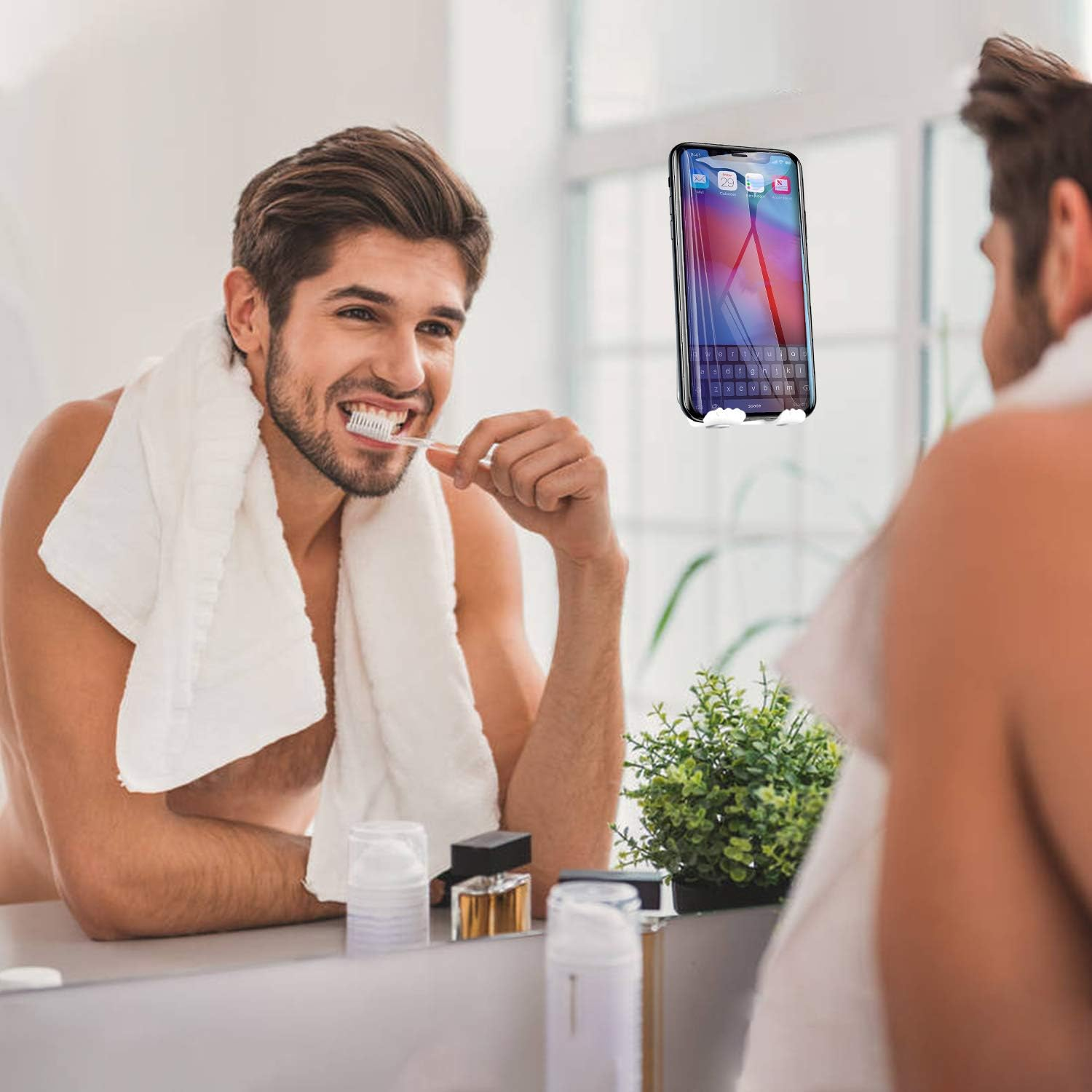 Ein Mann putzt sich vor einem Spiegel die Zähne. Ein Telefon ist am Spiegel befestigt. Ein Handtuch liegt um seinen Hals.