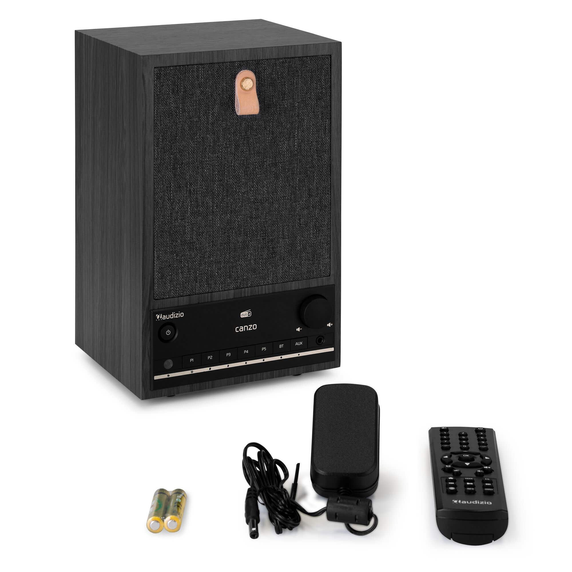 Un altavoz negro con mando a distancia, adaptador de corriente y pilas. El altavoz tiene una parte frontal de tela y un asa de cuero.