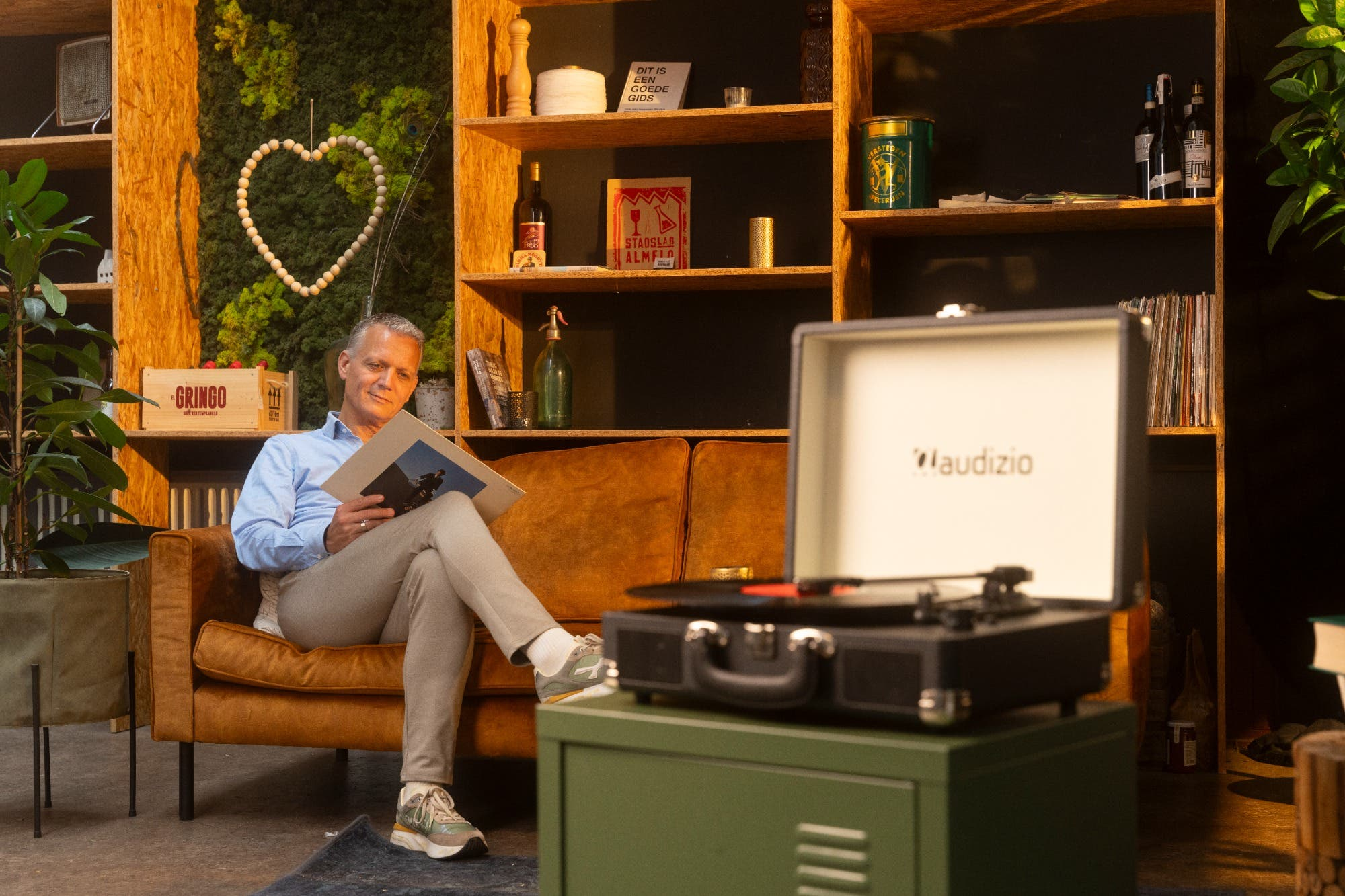 Un hombre sentado en un sofá marrón leyendo una portada de vinilo. Un tocadiscos sobre un armario verde. Estanterías con decoración detrás.