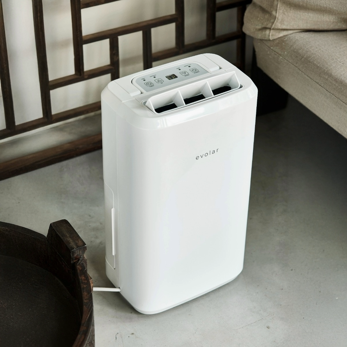 A white Evolar dehumidifier sits on a gray floor. A wooden screen and a couch are in the background.