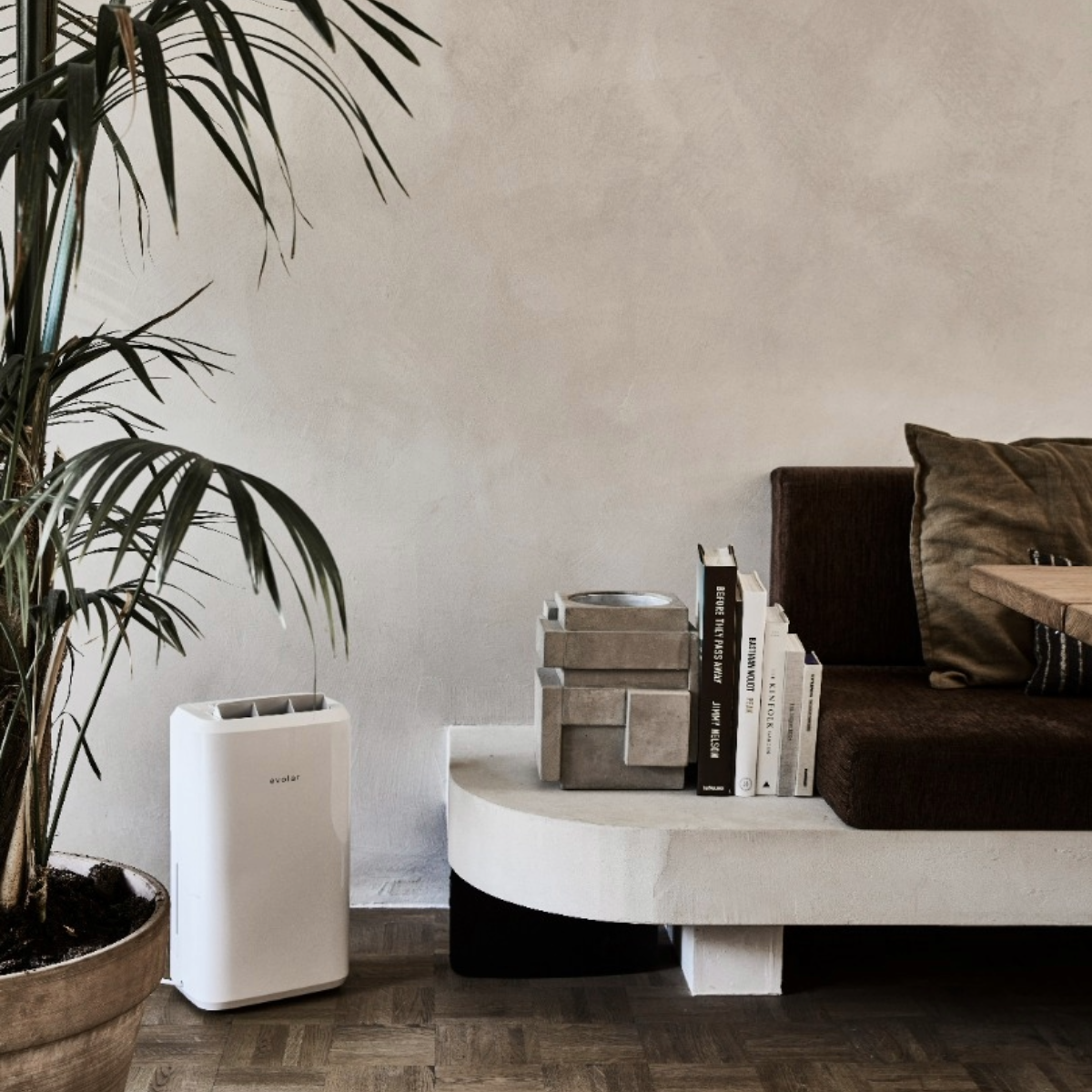 A white air purifier and green plant next to a brown sofa with books, a concrete sculpture, and a wooden table in a room.