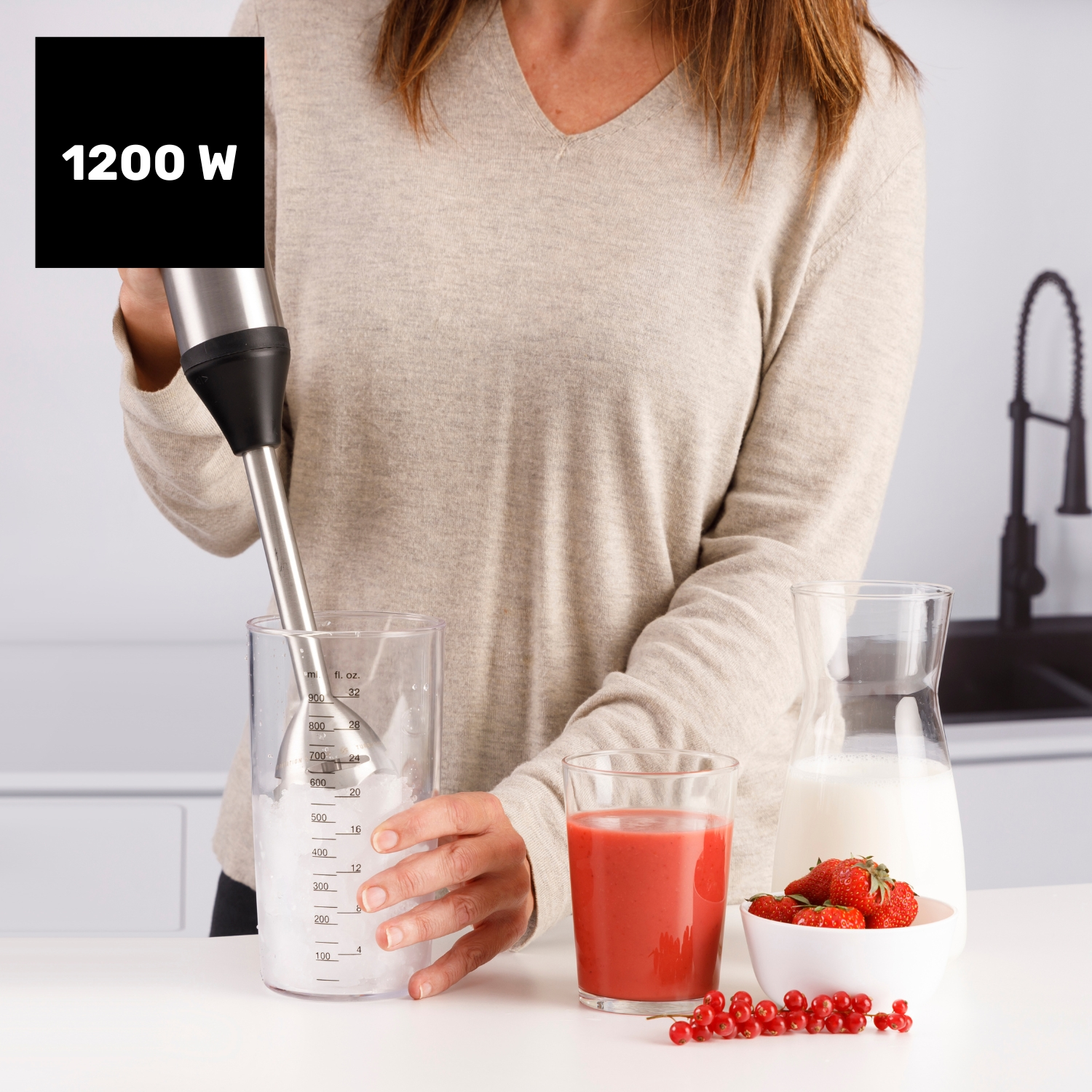 Una mujer usa una batidora. Los ingredientes incluyen hielo en una taza medidora, jugo rojo, fresas, leche y grosellas rojas.