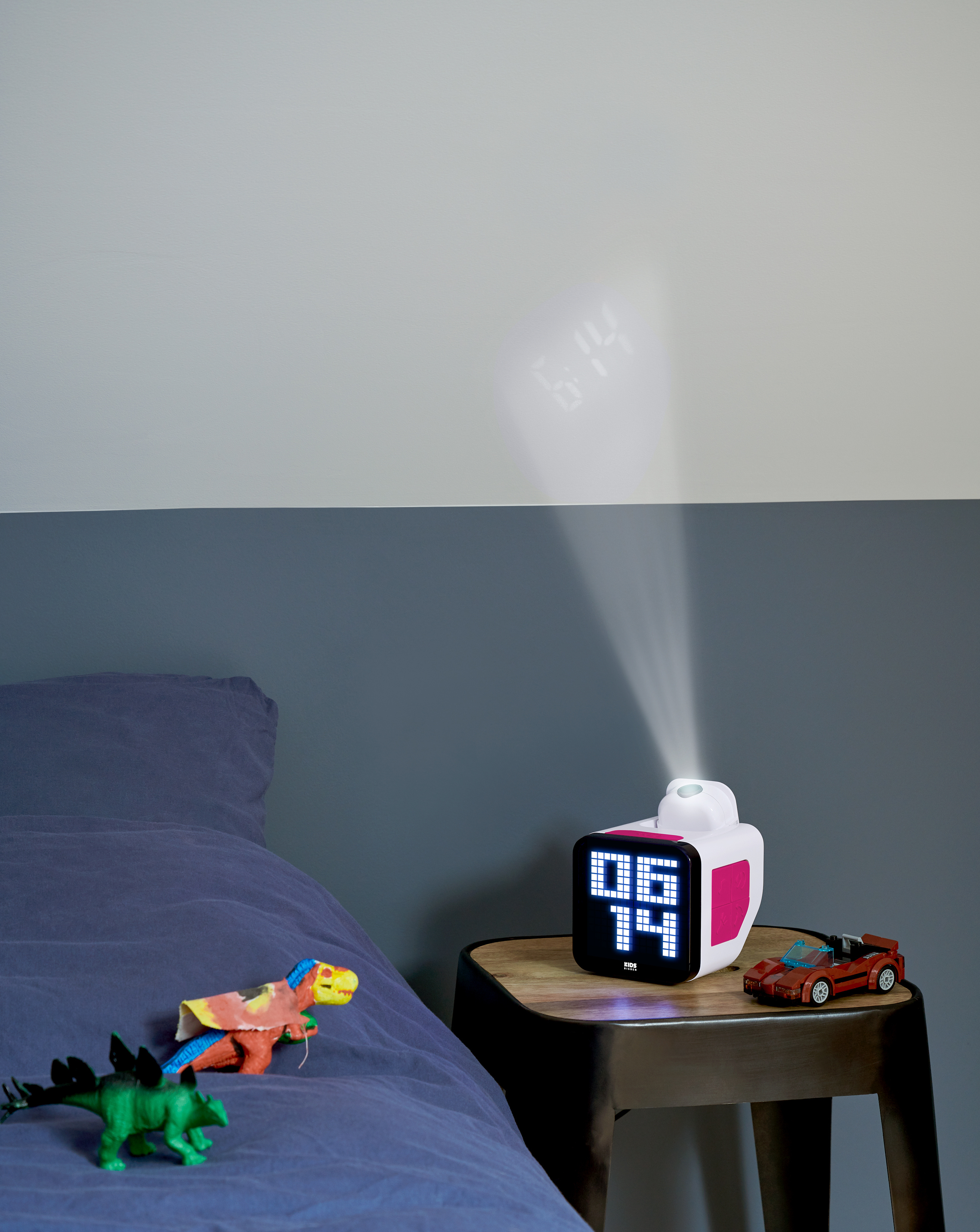 Una mesita de noche con un reloj digital, un coche de juguete y un haz de luz que proyecta la hora en la pared. Juguetes de dinosaurios en una cama.