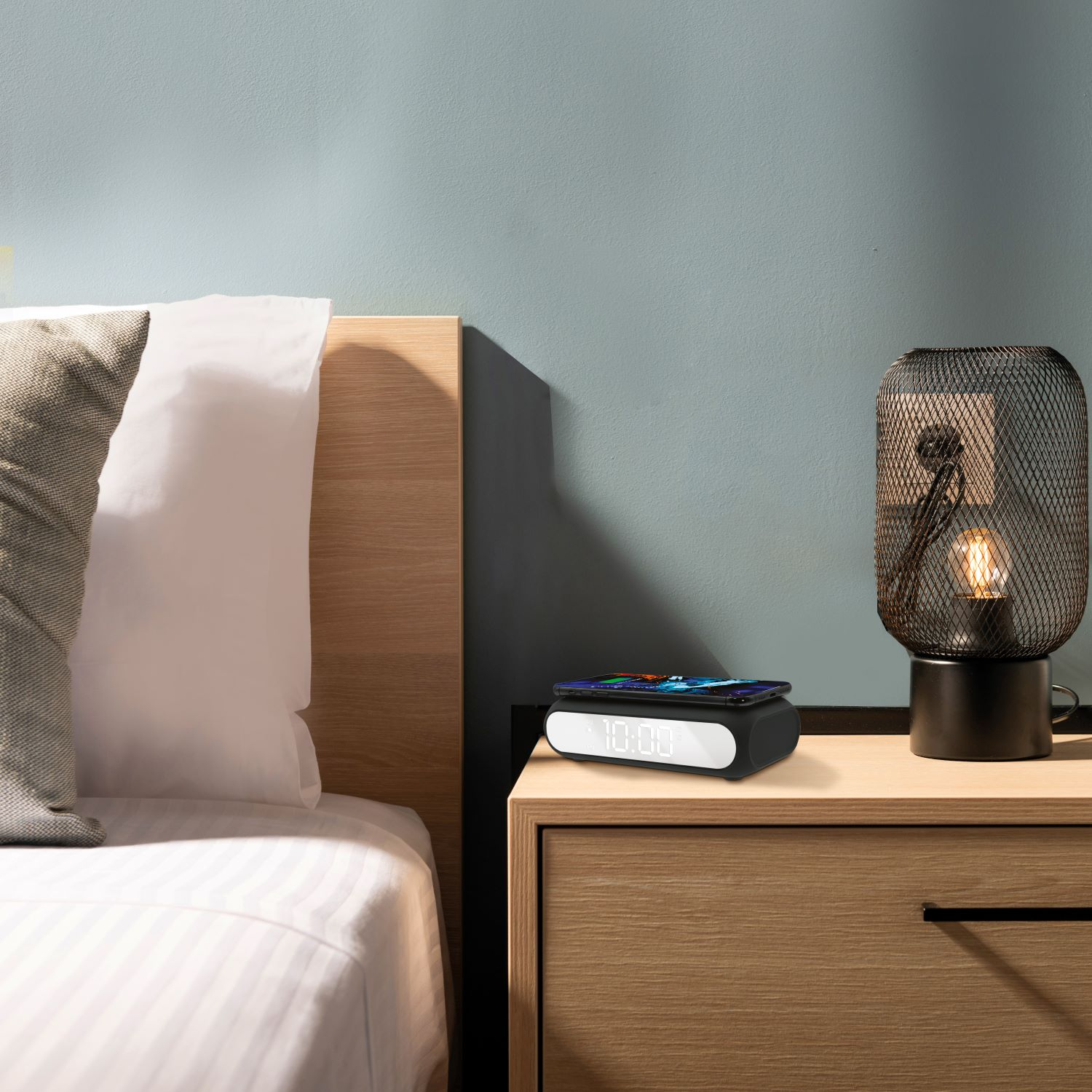 Escena de dormitorio con cama, mesita de noche, lámpara y reloj. El reloj muestra las 10:00. Un teléfono está en el reloj.