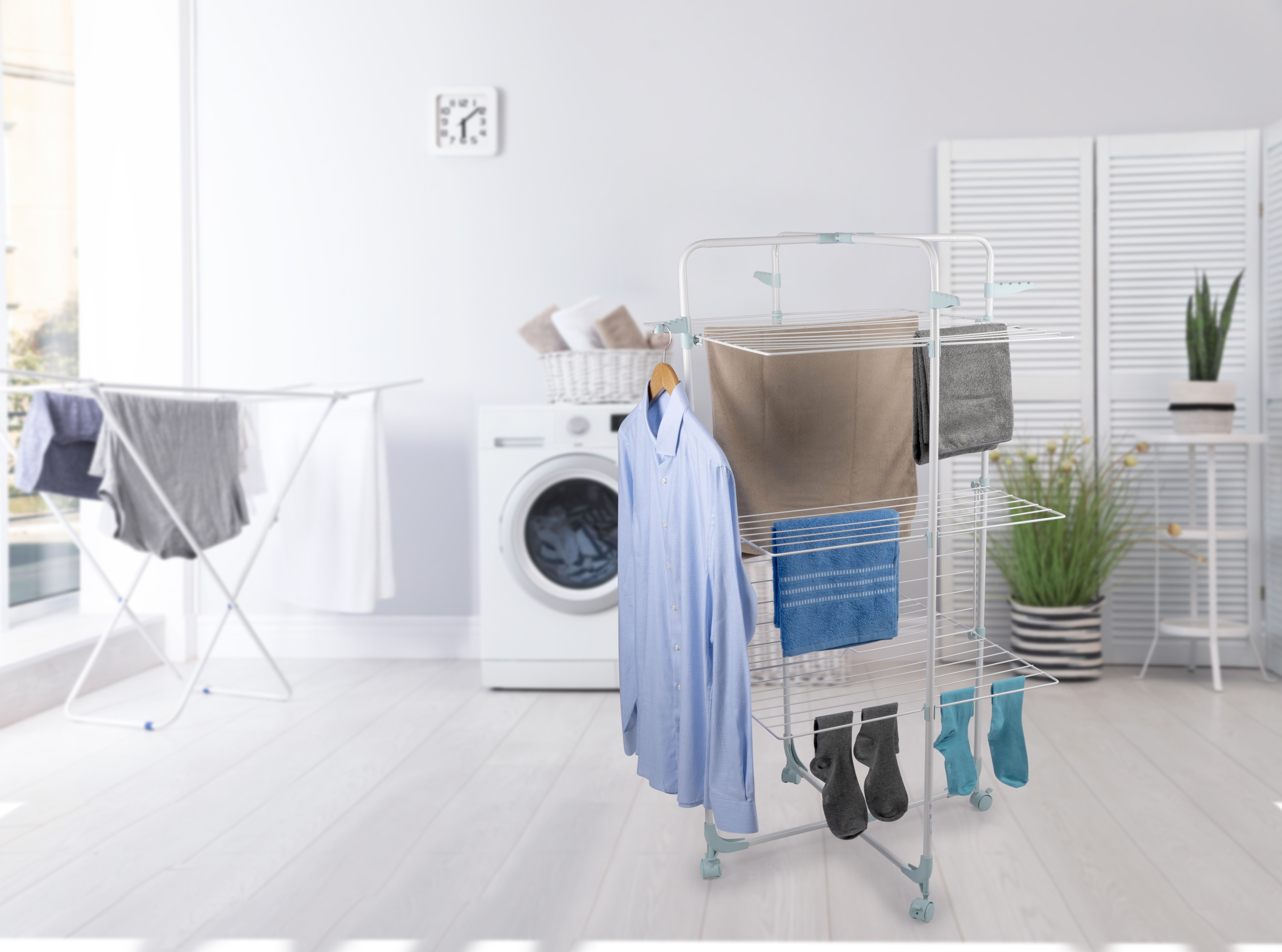 A laundry room with clothes drying on racks. A washing machine and a clock are visible. Plants and white screens in background.
