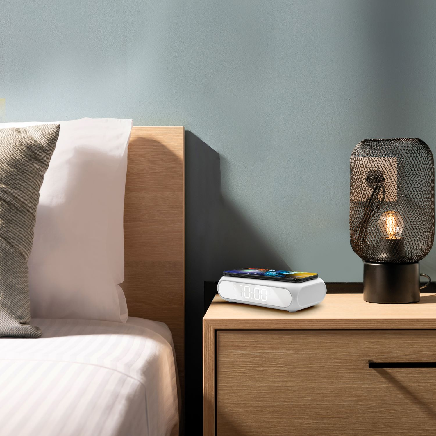Una mesita de noche con un reloj blanco y un teléfono encima. Una lámpara con base negra y una cama al fondo.