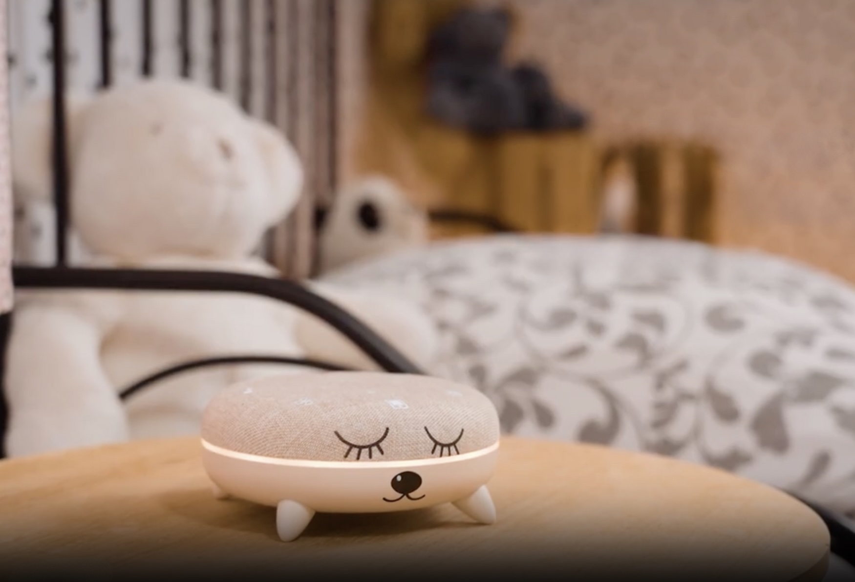 Un piccolo altoparlante bianco con una faccia è su un tavolo di legno. Un orsacchiotto e un letto sono sullo sfondo sfocato.