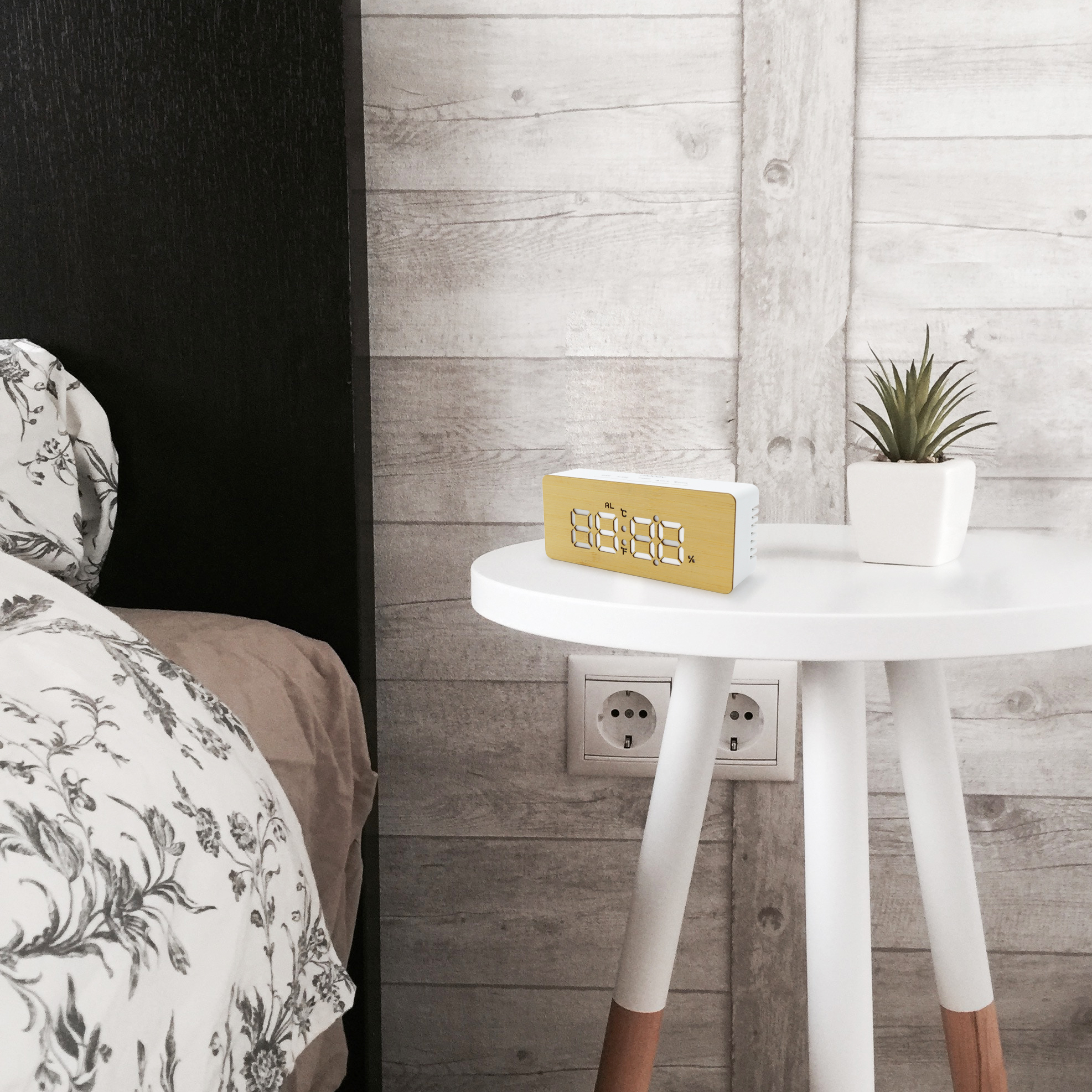Una escena de mesita de noche con una mesa blanca con reloj y planta. A la izquierda, una cama con sábanas estampadas.