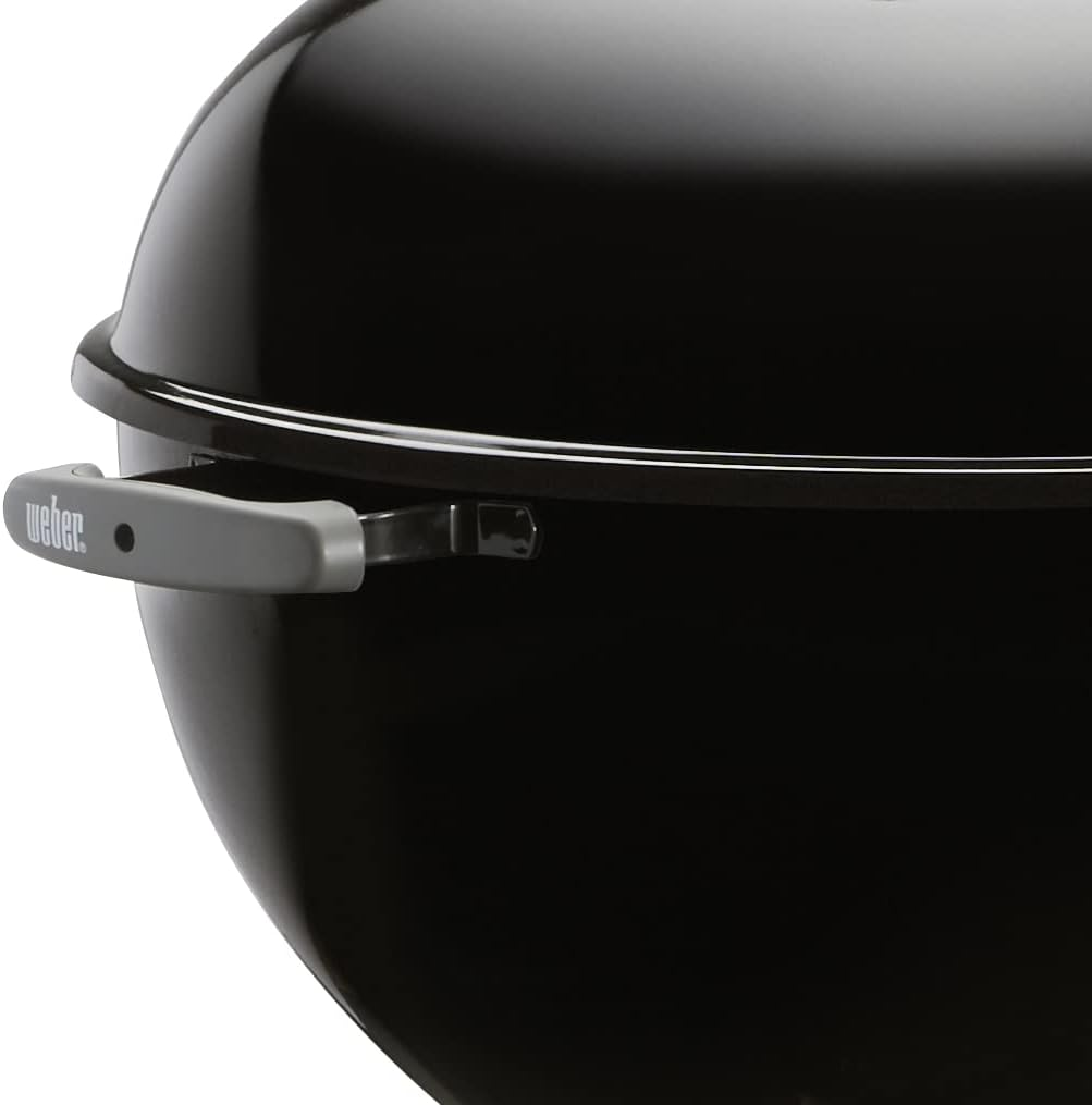 Primo piano di una griglia Weber nera. È visibile una maniglia grigia con il logo Weber. La superficie curva riflette la luce.