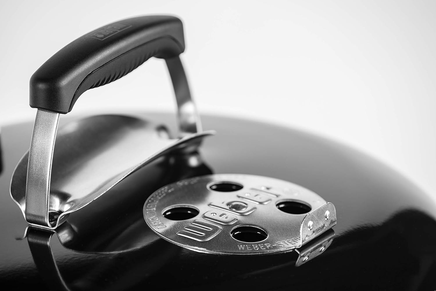 Primo piano di un coperchio nero per griglia con maniglia e ventilazione. La ventilazione è etichettata 'Weber-Step'.