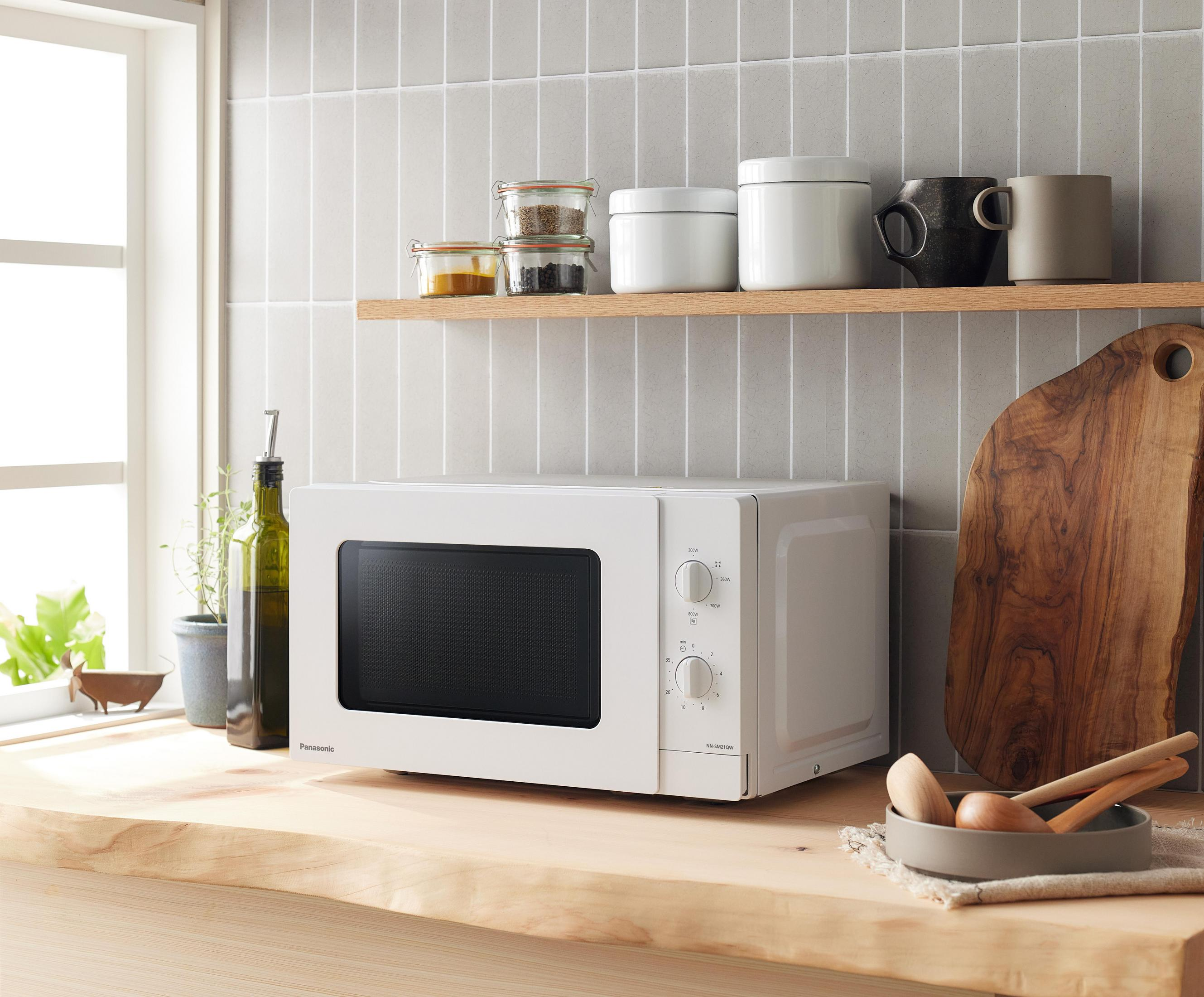 Biała kuchenka mikrofalowa na drewnianym blacie. Słoiki, kubki i deska do krojenia na półce. Okno po lewej.