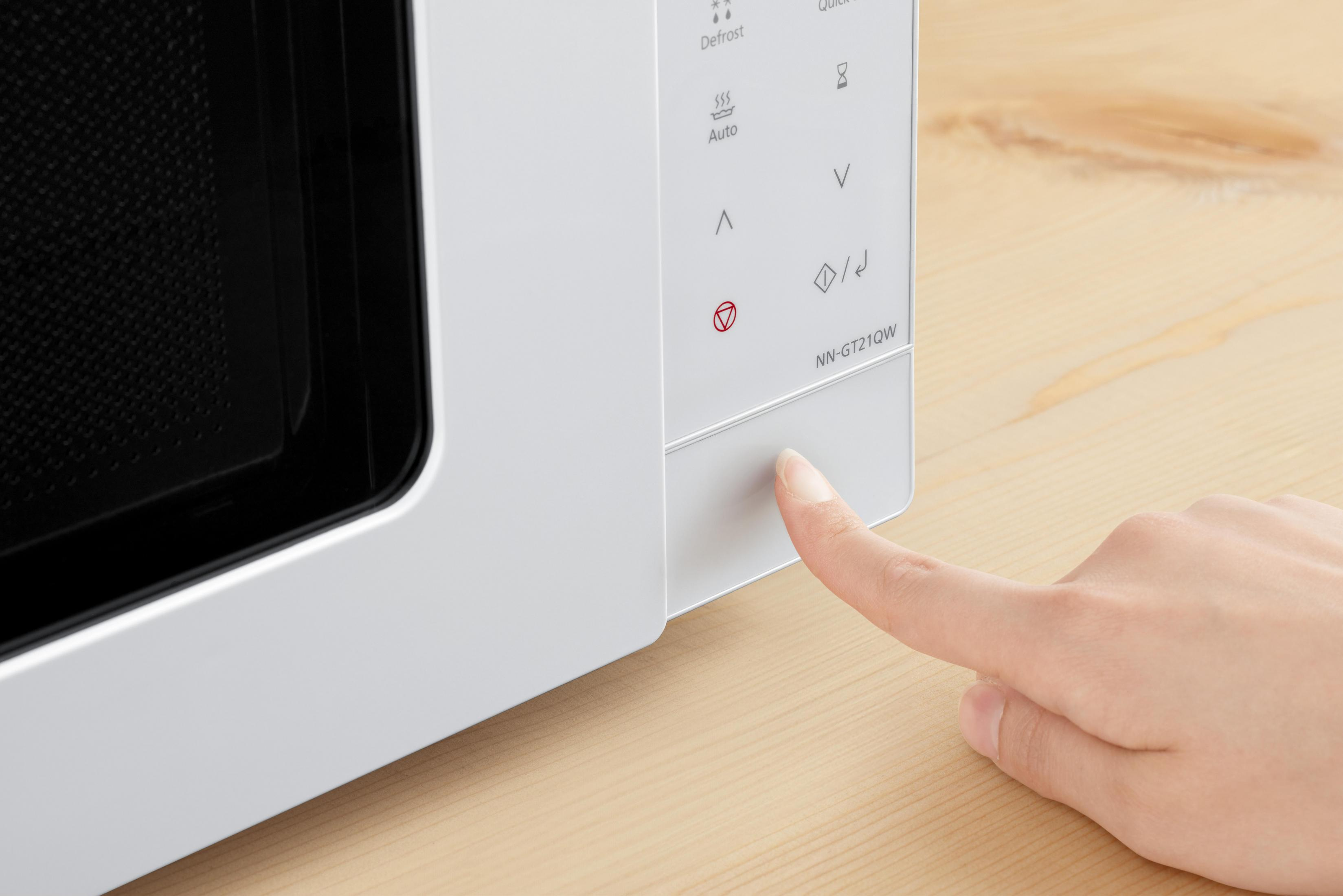 Una mano preme un pulsante su un forno a microonde bianco. Il microonde è su una superficie di legno chiaro.