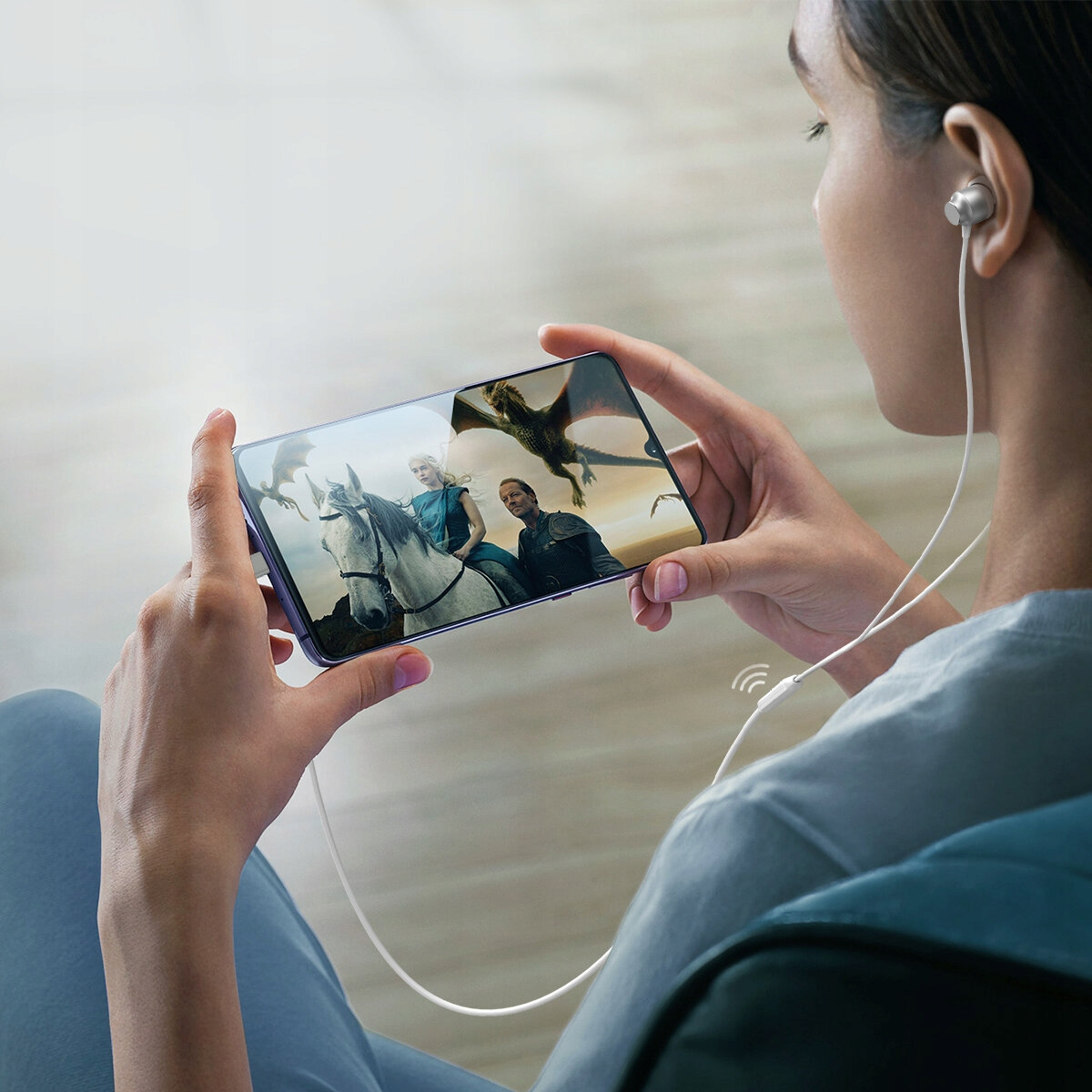 Osoba ogląda program na telefonie, mając słuchawki. Telefon wyświetla scenę ze smokami i ludźmi na koniach.