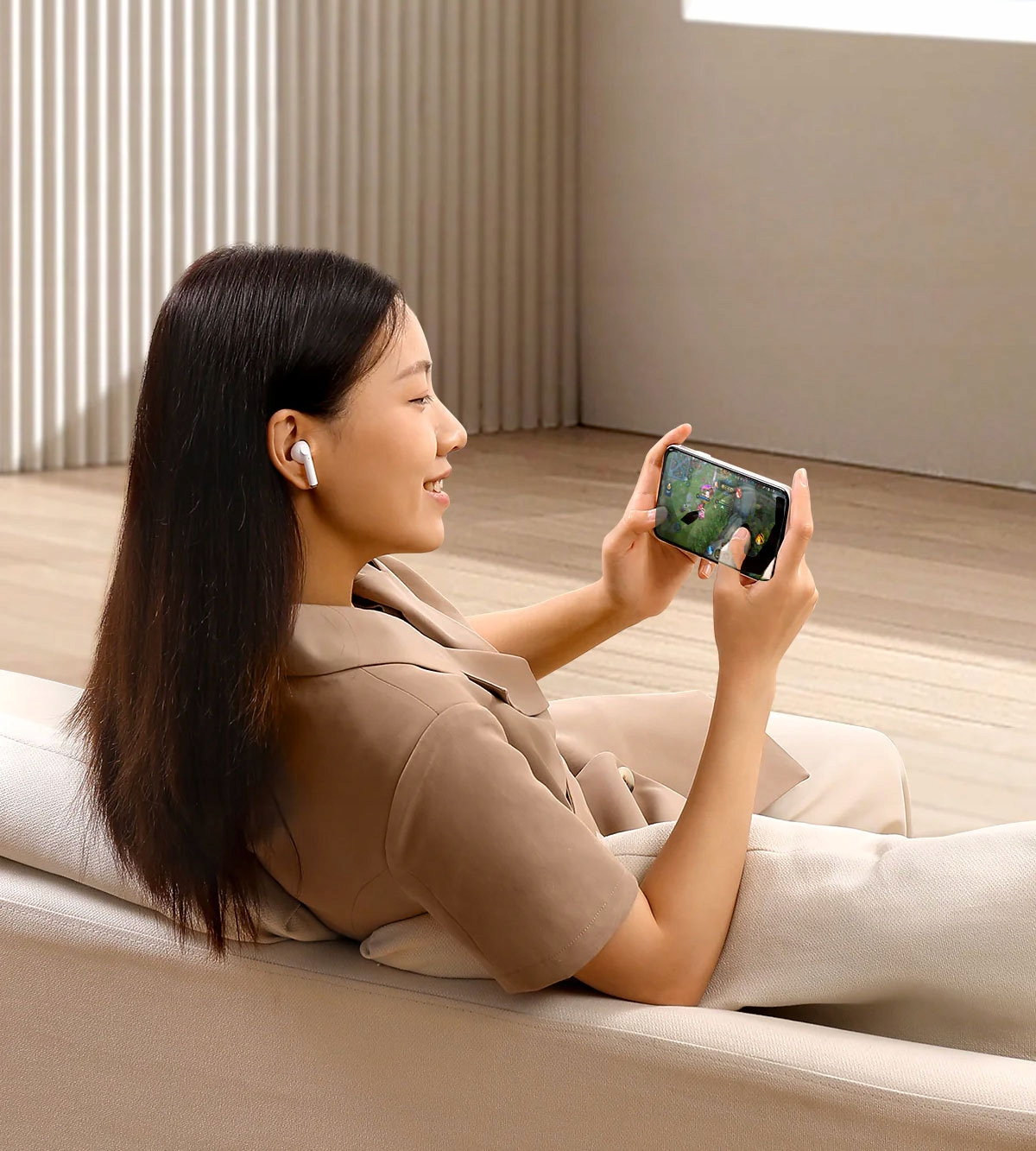 Una donna è seduta su un divano, indossa auricolari e gioca sul suo telefono. Sorride. Abiti e sfondo beige.