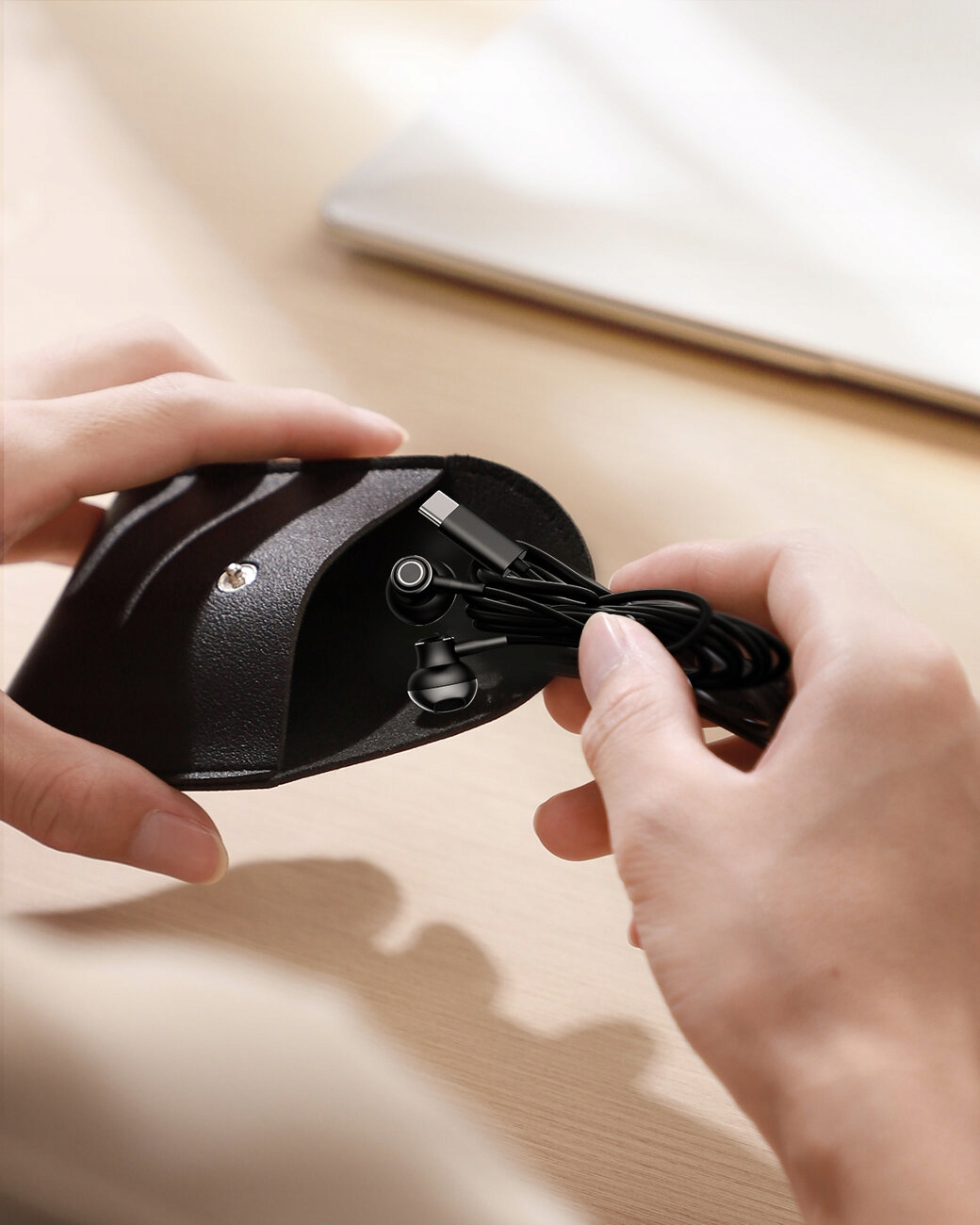 Ręce trzymają czarne słuchawki i etui. Laptop w tle. Słuchawki są podłączone i gotowe do użycia.