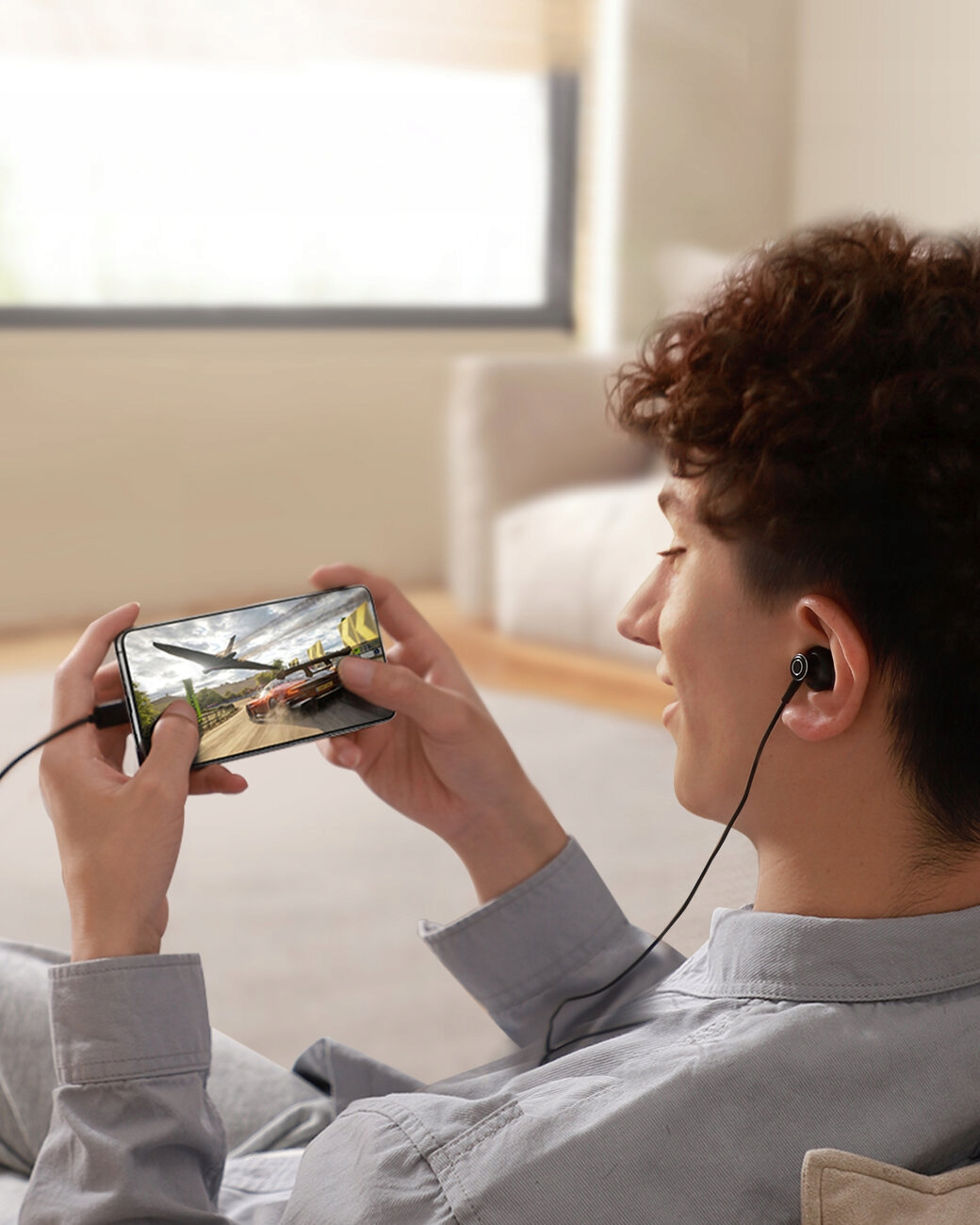 Osoba trzyma smartfon z grą wyścigową. Ma słuchawki i się uśmiecha. Tło obejmuje okno i sofę.