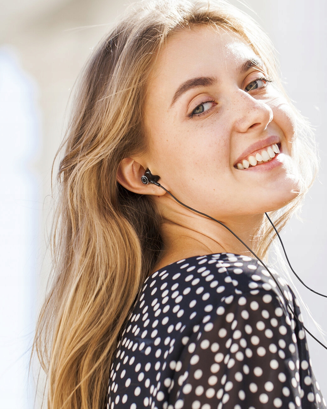 Blondynka uśmiecha się, nosi słuchawki i sukienkę w kropki. Spogląda w stronę kamery, z miękkim światłem słonecznym w tle.
