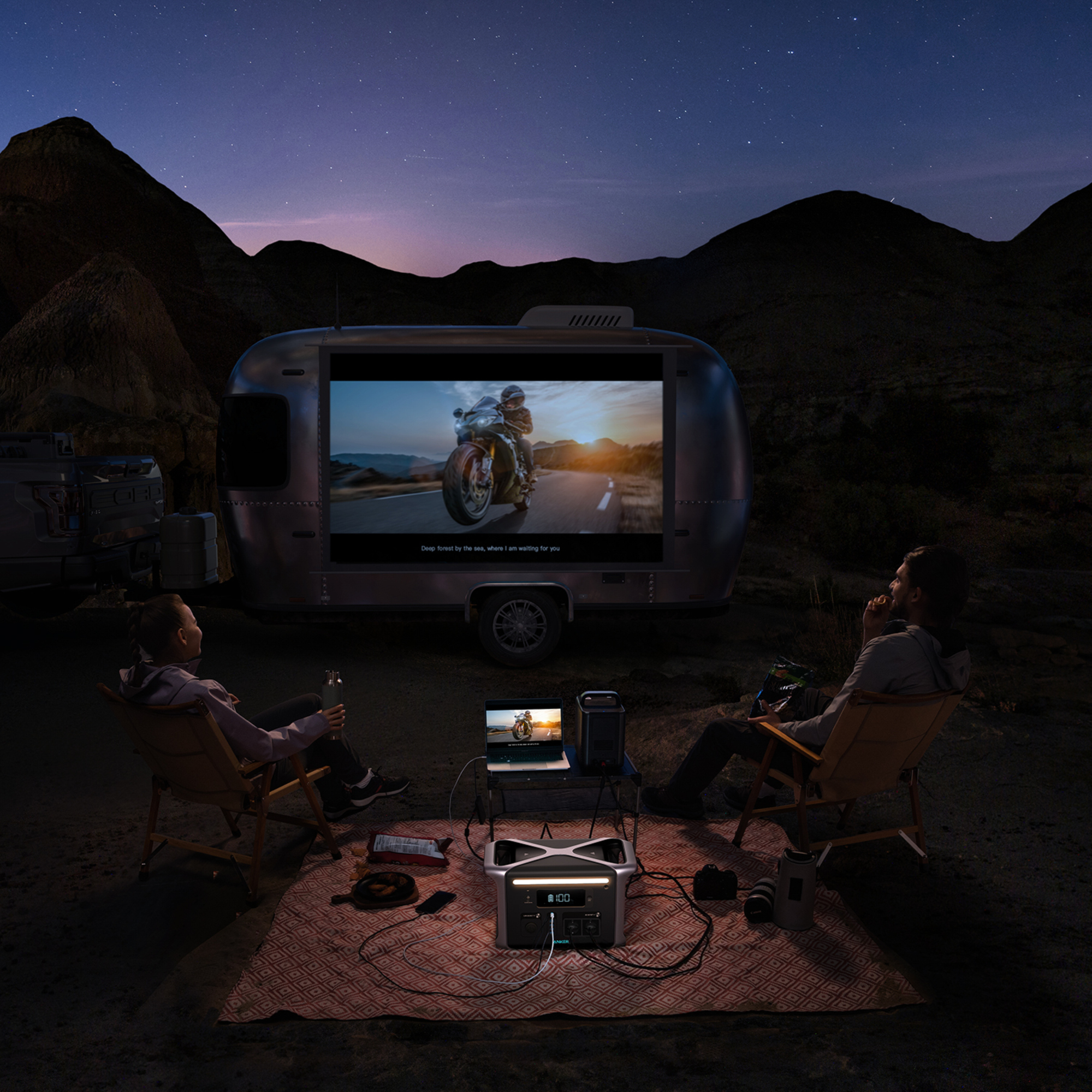Dwie osoby oglądają film na ekranie przyczepy w nocy. W tle góry i gwiazdy. Akcesoria na pierwszym planie.