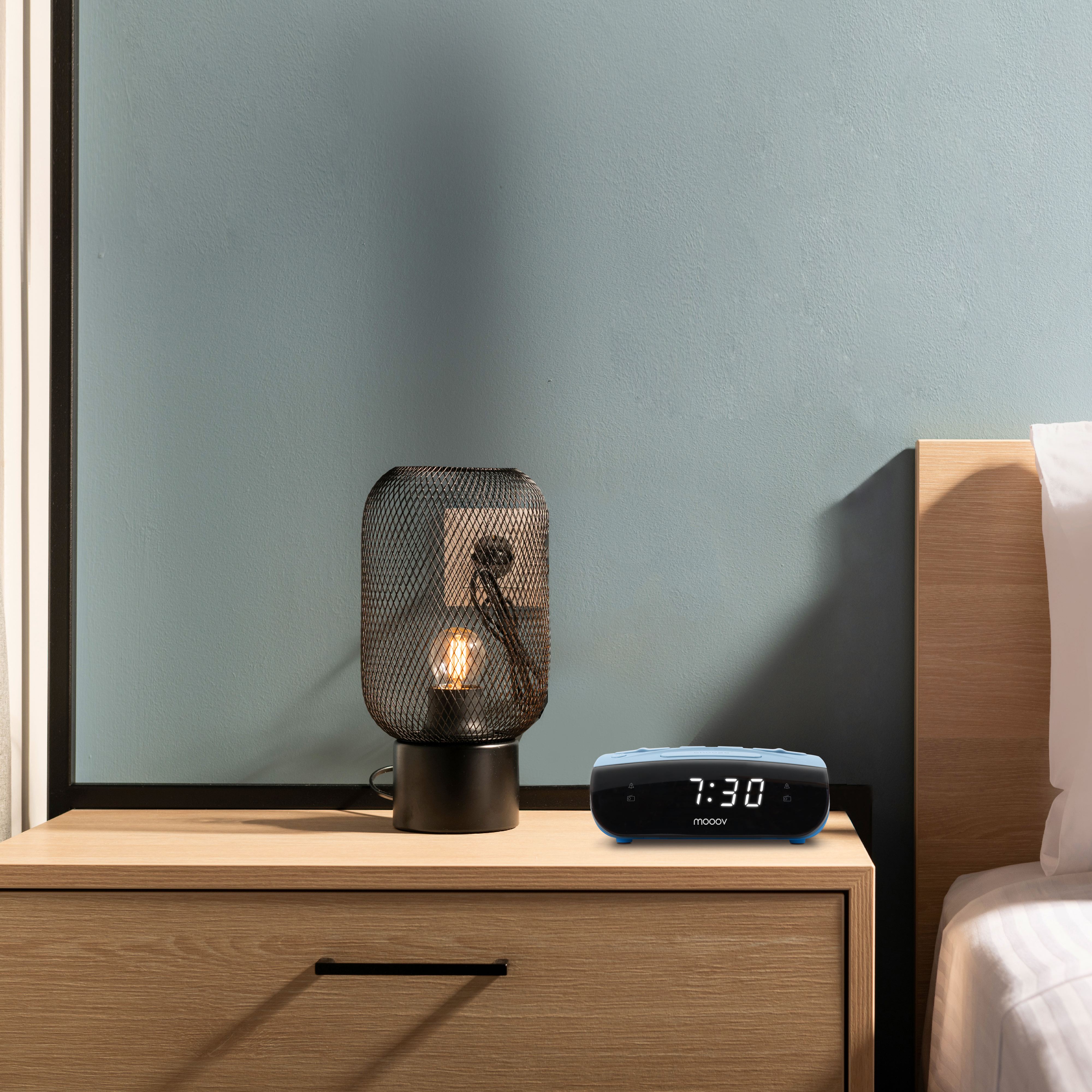 Una mesita de noche con una lámpara y un despertador. El fondo es una pared azul y parte de una cama.