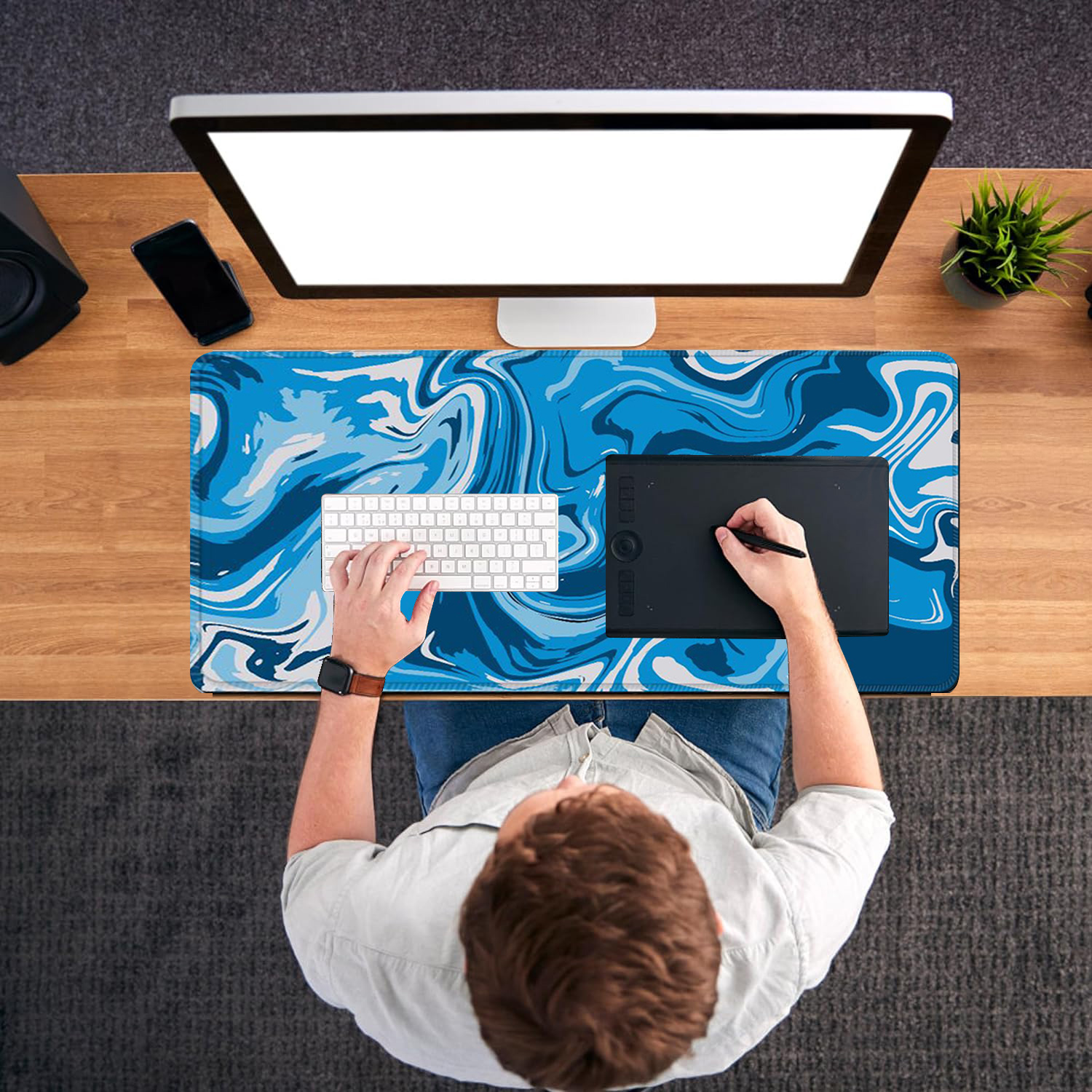 A person is working at a desk with a computer. A blue and white patterned desk mat, keyboard, and drawing tablet are visible.
