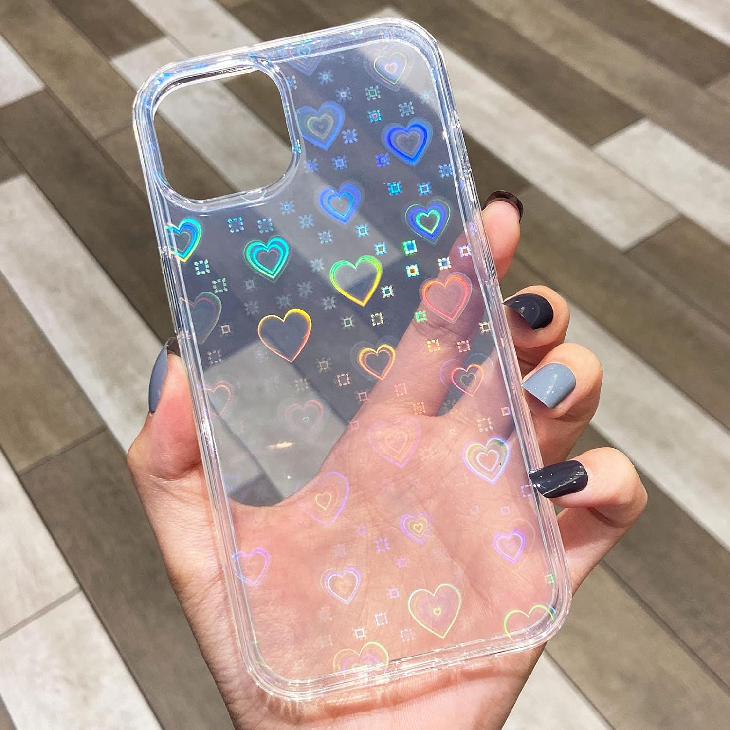 Eine Hand hält eine transparente Handyhülle mit holografischen Herzen. Der Hintergrund ist ein Holzboden. Die Nägel sind in verschiedenen Farben lackiert.