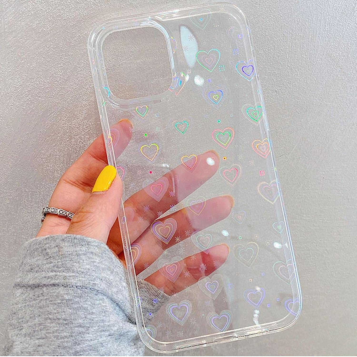 Hand hält transparente Handyhülle mit Herzmustern. Gelber Nagellack, Silberring, grauer Pullover. Der Hintergrund ist hellgrau.