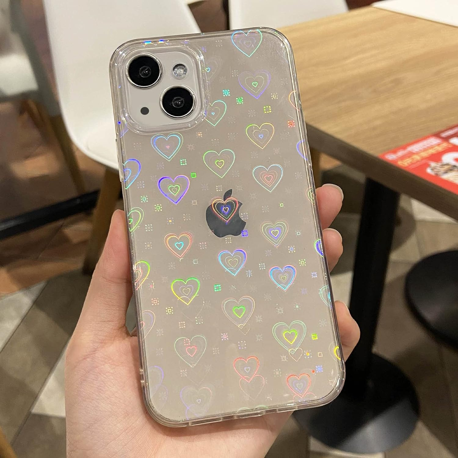 Eine Hand, die eine durchsichtige Handyhülle mit Herzen hält. Die Hülle befindet sich auf einem iPhone mit schwarzem Apple-Logo und Dual-Kamera.