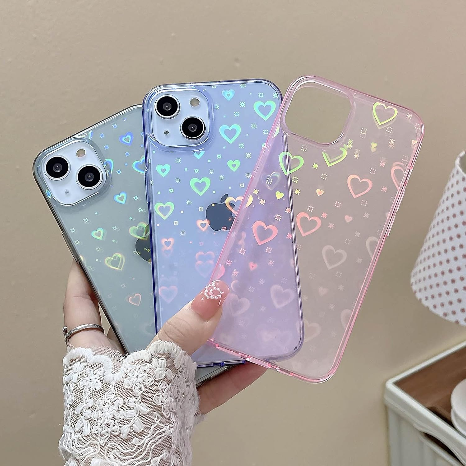 Hand hält drei iPhone-Hüllen. Die Hüllen sind blau, lila und rosa und mit Herzen verziert. Die Hand trägt einen Spitzensaum und einen Ring.