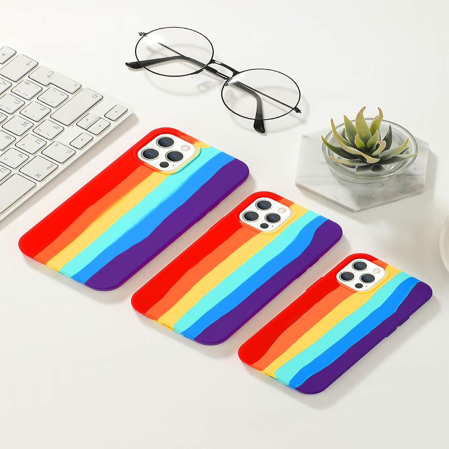 Drei Regenbogen-Handyhüllen auf einem weißen Schreibtisch mit Tastatur, Brille und einer kleinen Pflanze.